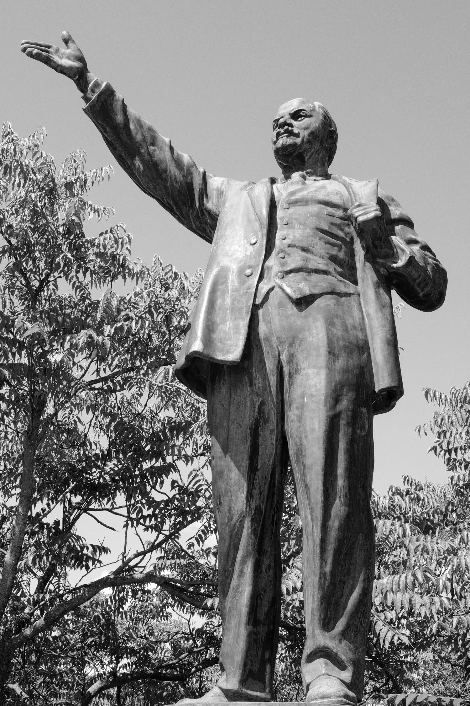 statue en bronze d'un homme debout, pointant vers l'avant avec sa main droite, portant un veston et une cravate, entouré d'arbres