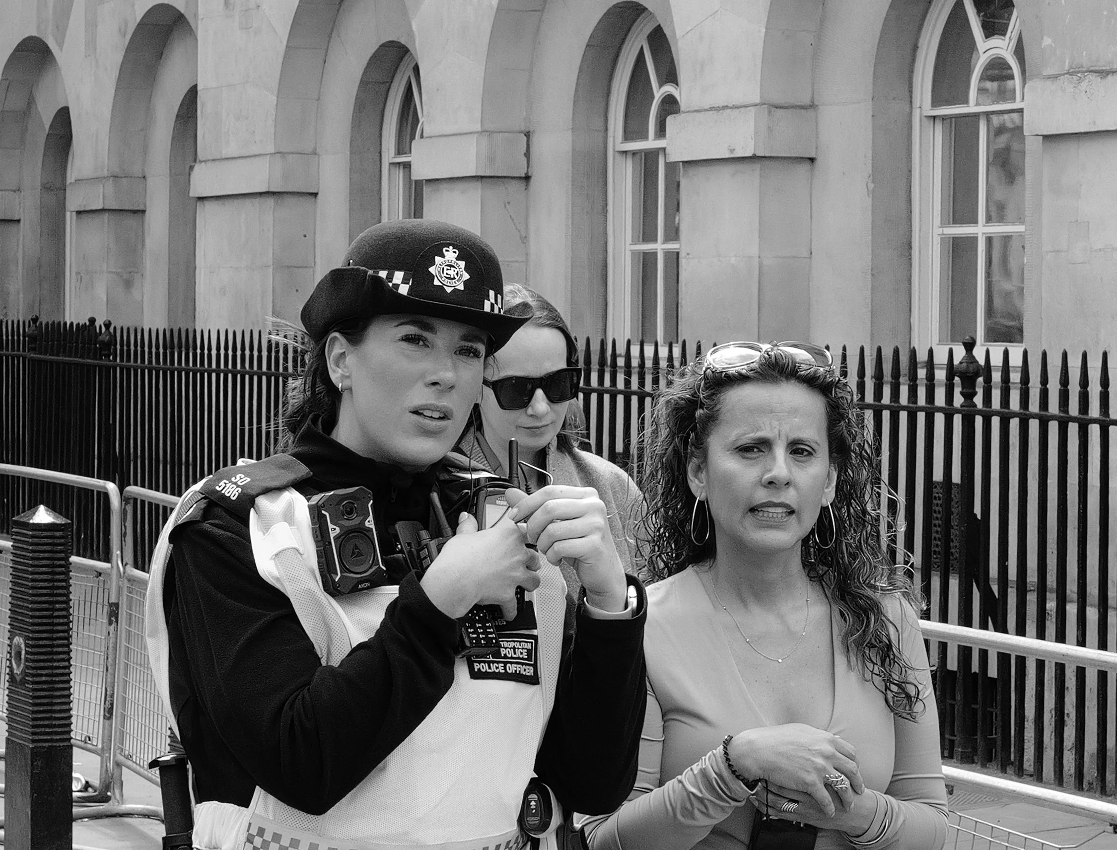 Trois femmes, dont une policière en uniforme, posent pour une photo devant un bâtiment en pierre avec des fenêtres arquées. La policière porte un casque et un gilet pare-balles, une autre femme porte des lunettes de soleil et une autre a les cheveux 