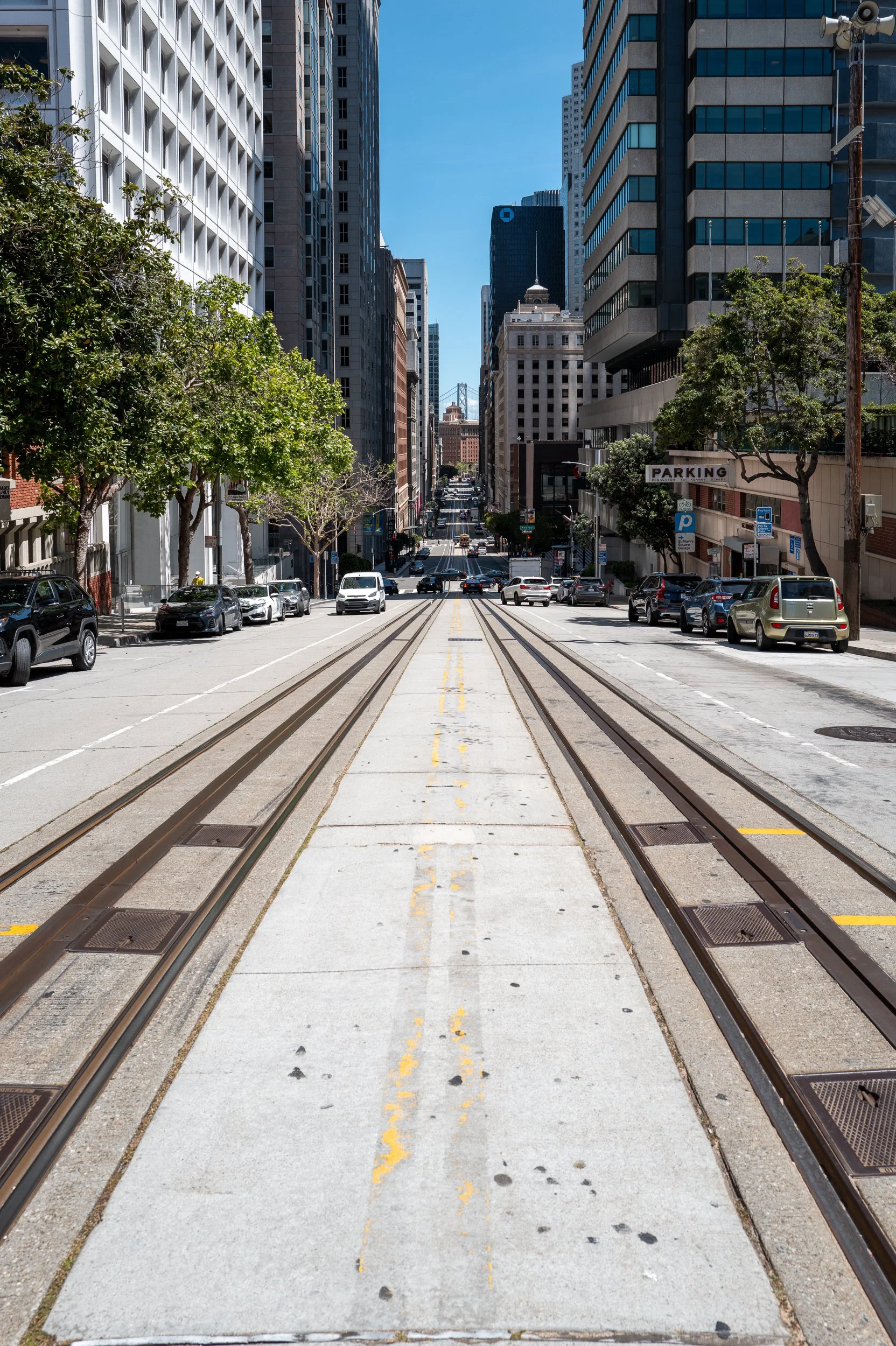 Rue urbaine avec tramway, bâtiments modernes et stationnement, arbres, voitures en stationnement, ciel clair.