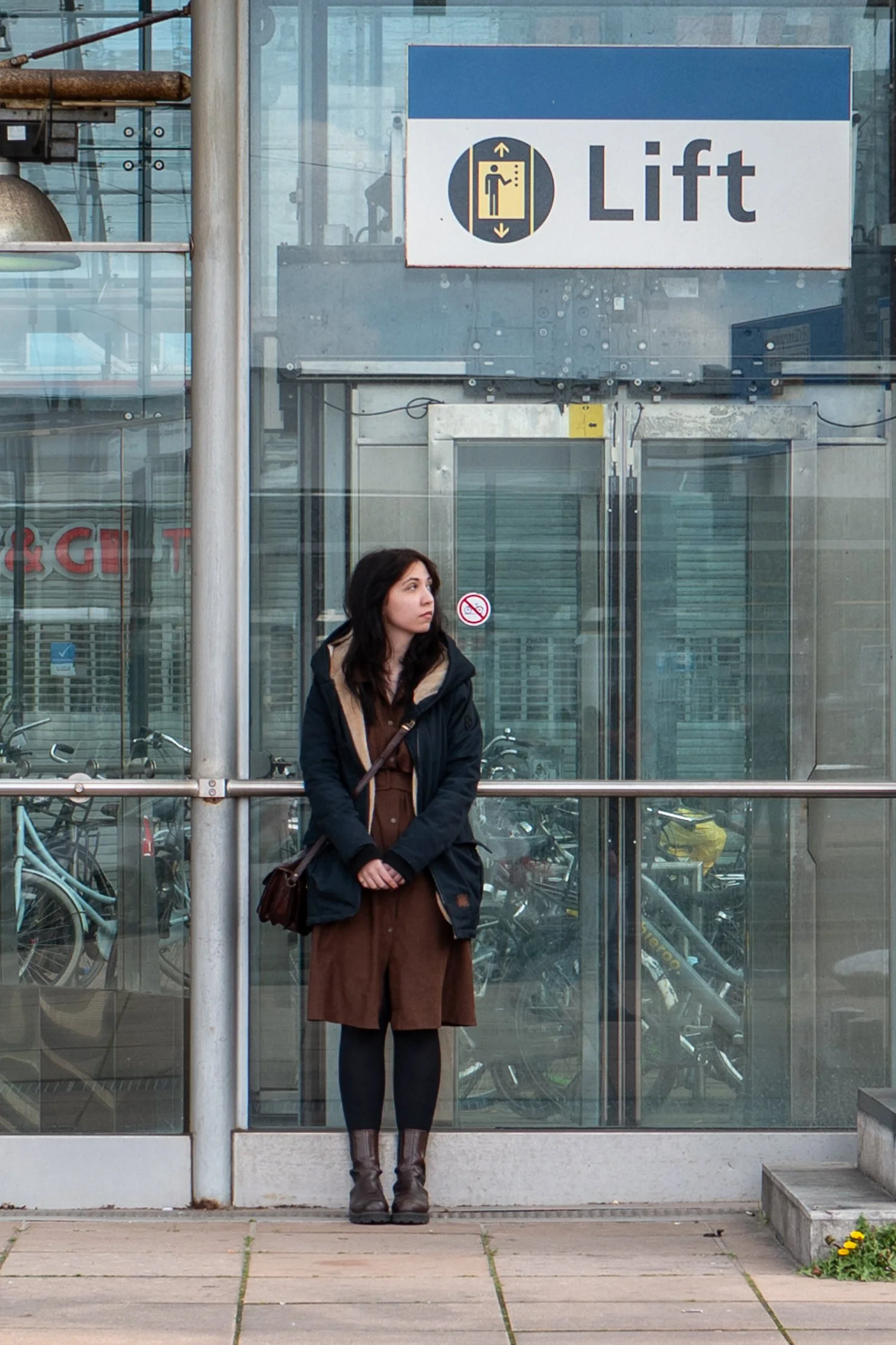 Une femme debout devant une porte d'ascenseur en verre, avec un panneau indiquant 'Lift' et un symbole d'ascenseur. Elle porte un manteau noir, une robe marron, des bottes marron, et a un sac en bandoulière. Il y a des vélos derrière la vitre et des 