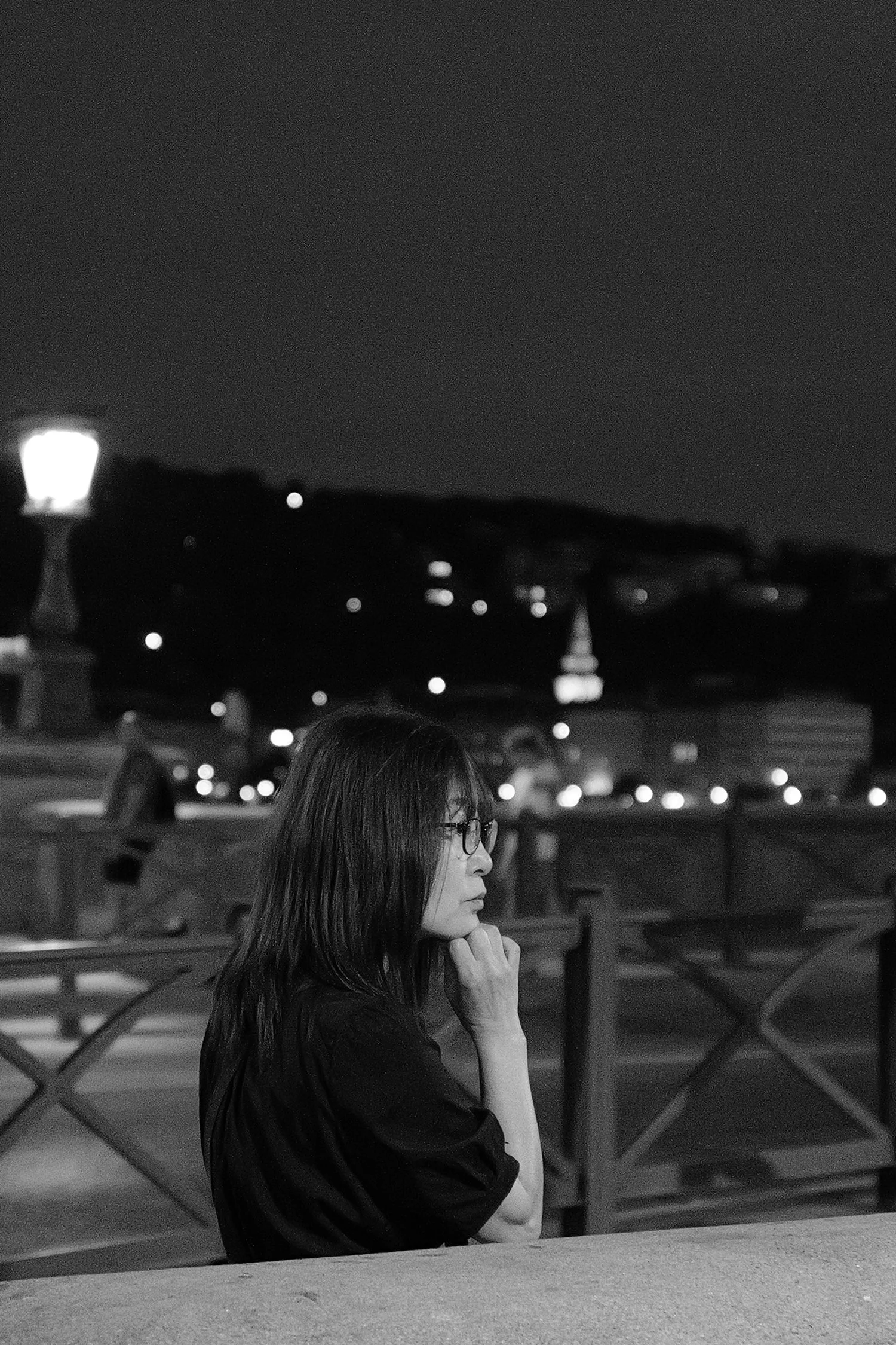 Une jeune femme aux lunettes, en profil, assise sur un banc, dans un lieu urbain la nuit, avec des lampadaires et des voitures en arrière-plan.