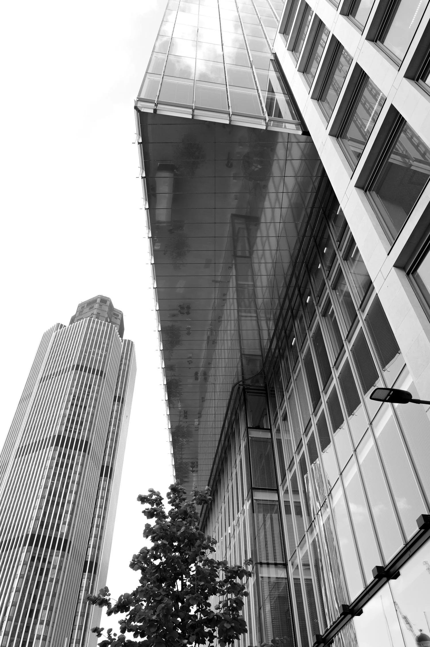 Vue d'un bâtiment moderne en verre avec un autre gratte-ciel dans la ville au fond, vu de bas en haut, et quelques arbres devant.