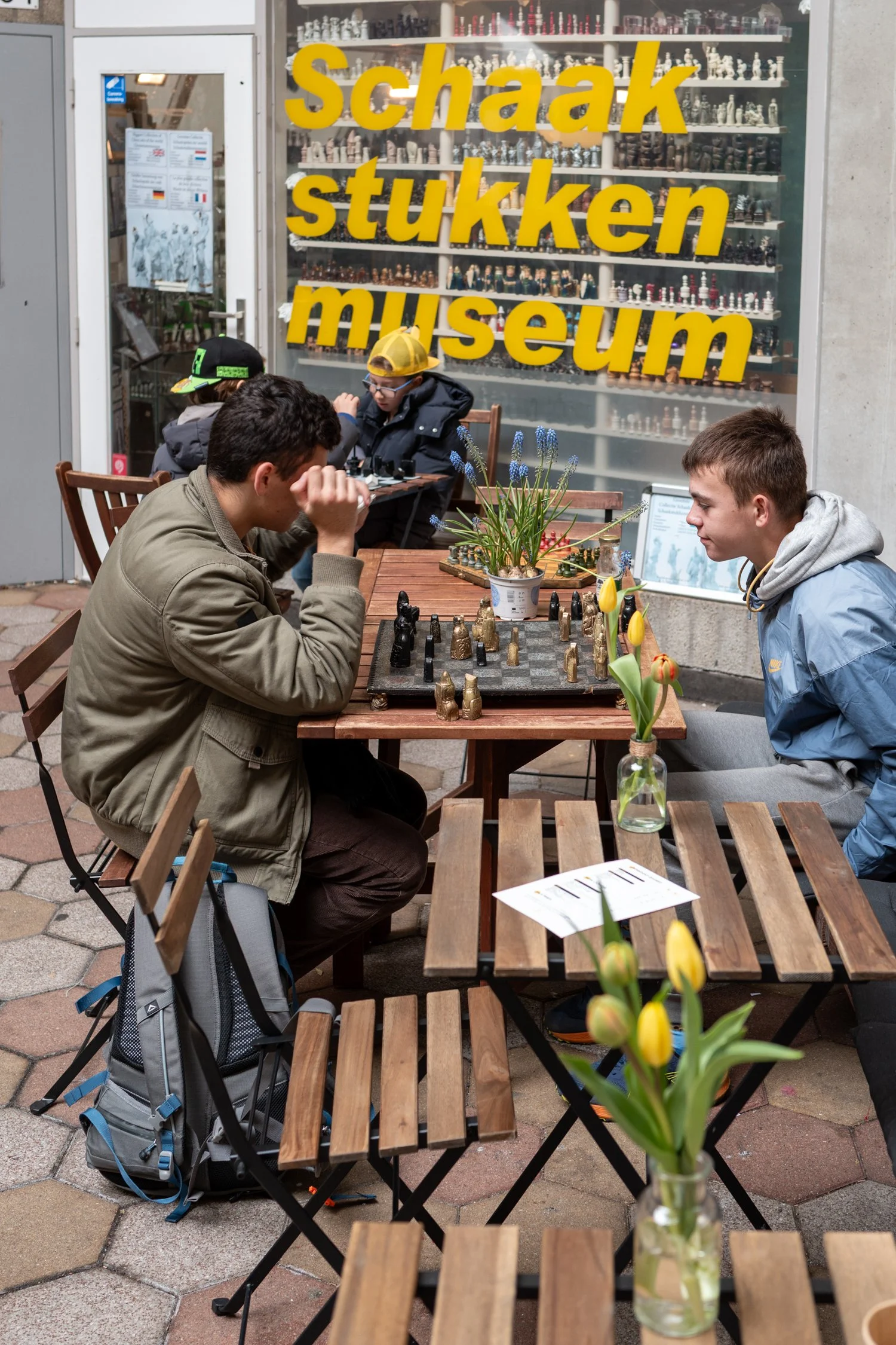 Des jeunes jouent aux échecs lors d'une activité en plein air devant un magasin de pièces d'échecs avec la vitrine en arrière-plan, où l'on peut lire 'Schaak stukken museum'.