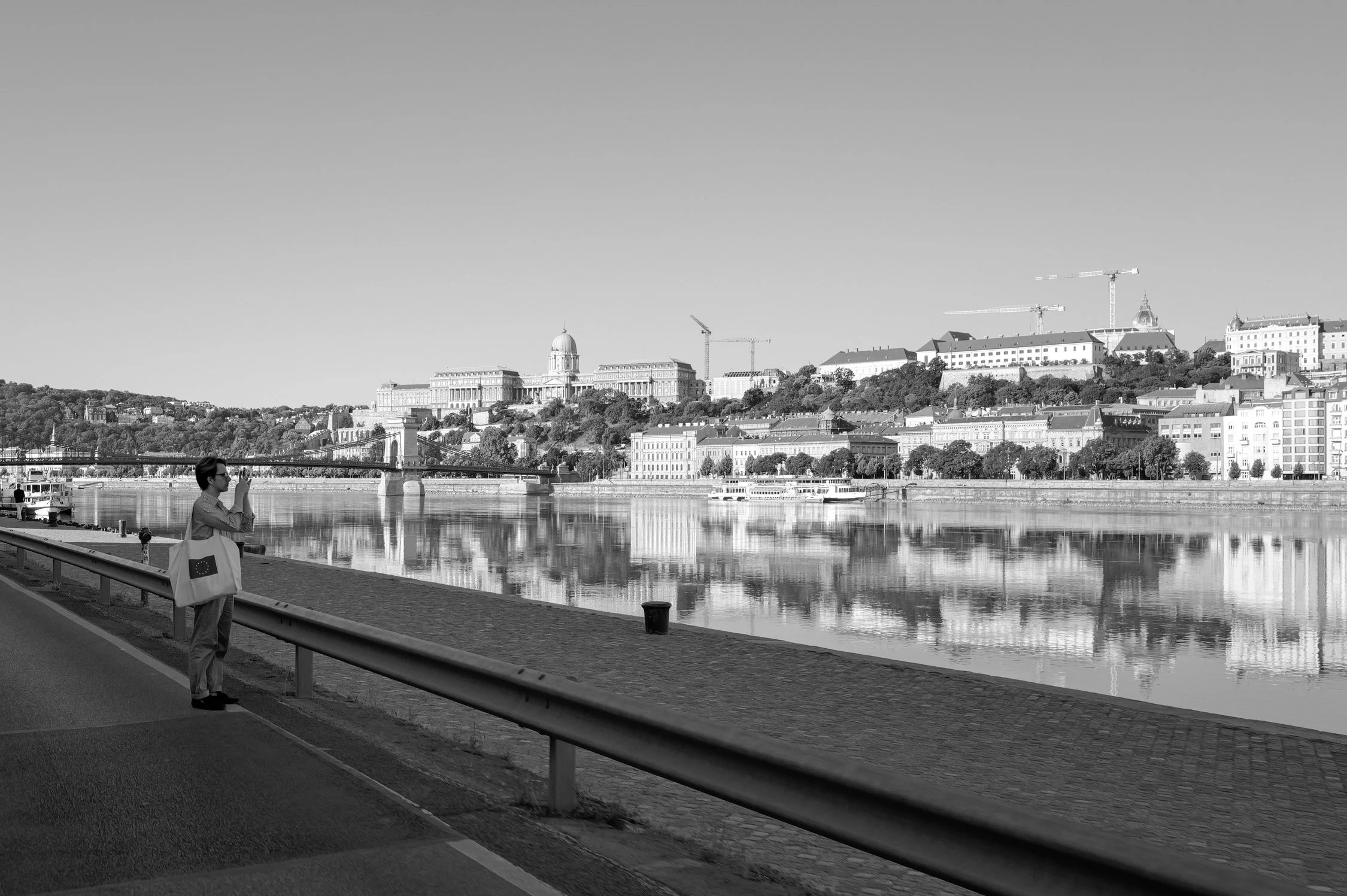 Une personne en costume se tient près de la rivière Szigetvár, avec une vue de la ville de Budapest et le Parlement hongrois en arrière-plan.