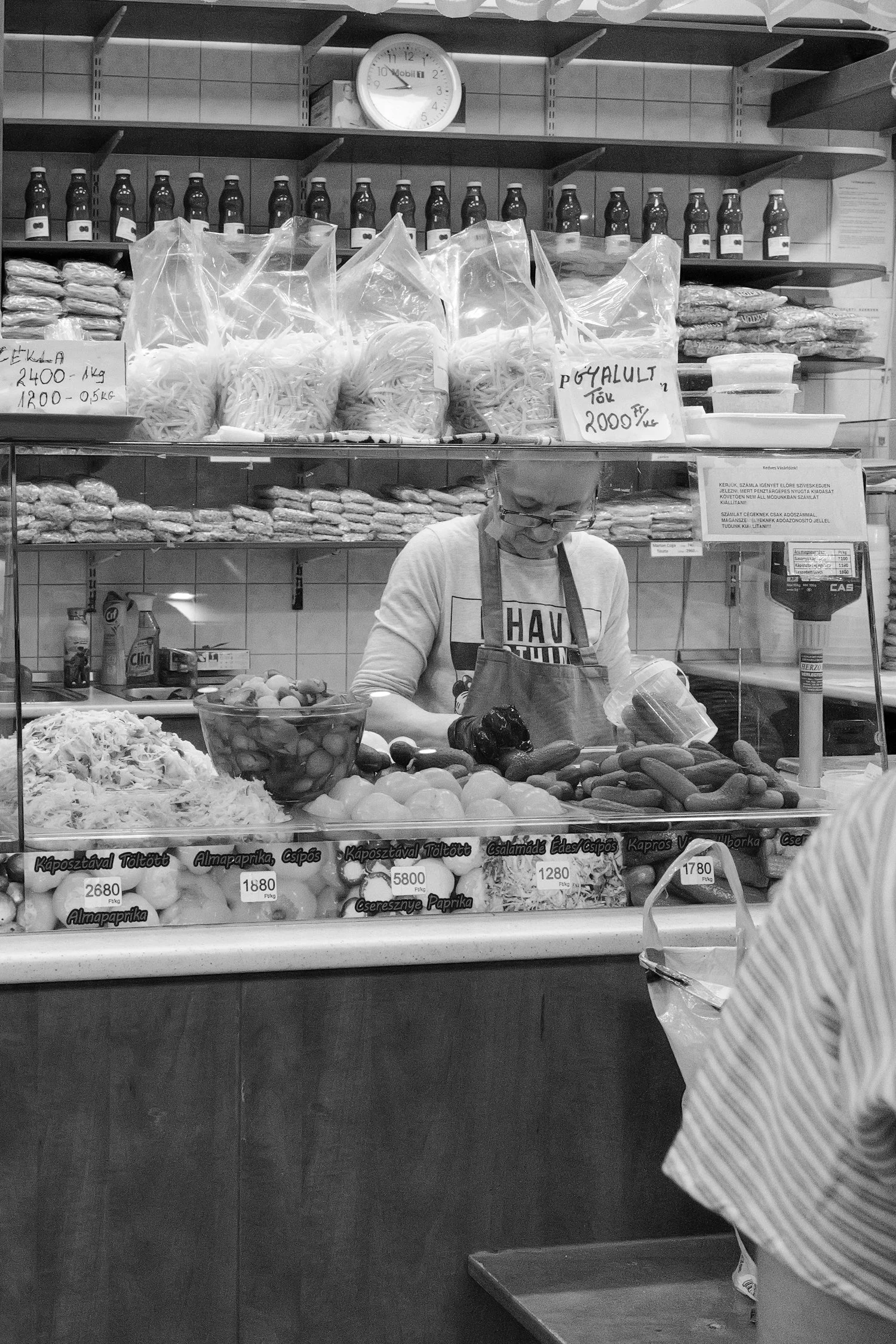 Vitrine de marché avec des légumes et des produits emballés, un employé prépare des articles, tableau de prix, et étagères remplies de bouteilles et de sacs.