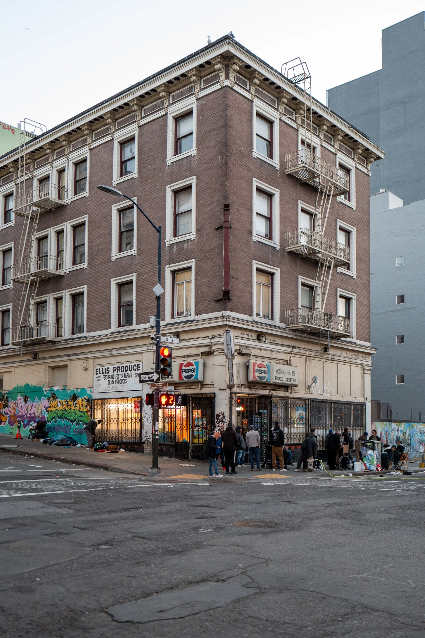 Un bâtiment en brique avec des escaliers d'urgence à l'extérieur, un restaurant ou un supermarché au rez-de-chaussée, et des gens faisant la queue ou rassemblés devant. Graffiti sur le mur à gauche, signal de feu rouge, rue urbaine avec sol usé.