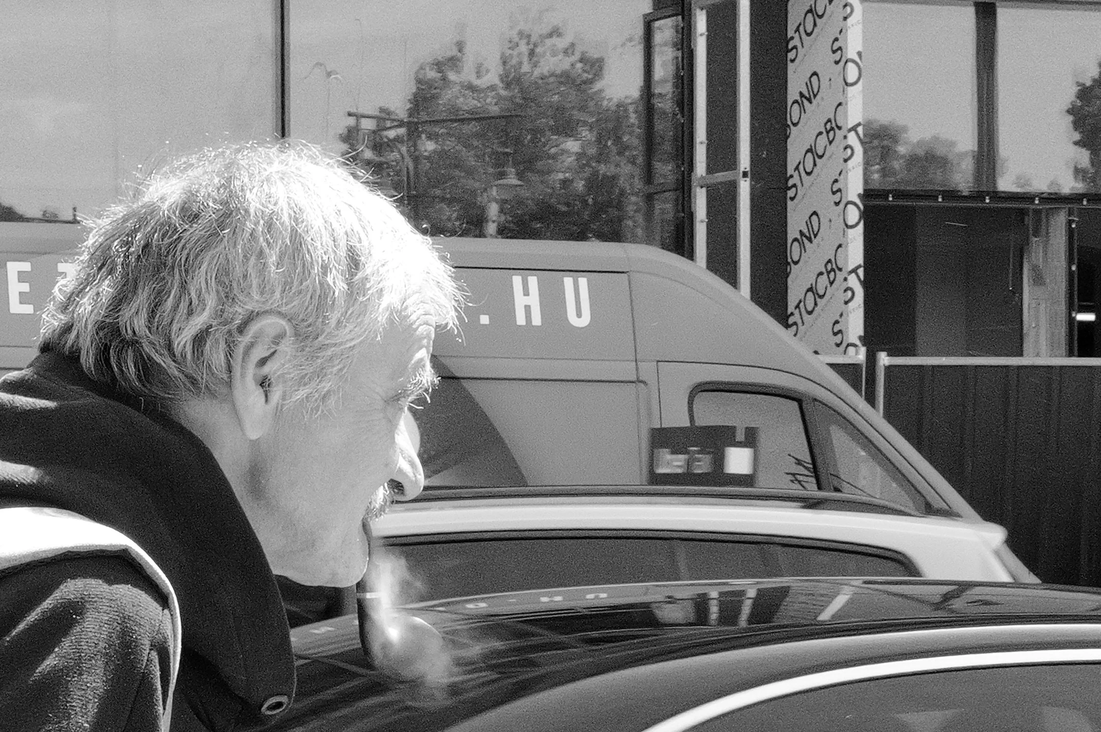 Une personne à cheveux gris à l'intérieur d'une voiture, vue de profil, avec un bâtiment en arrière-plan et une grande boîte ou panneau publicitaire.