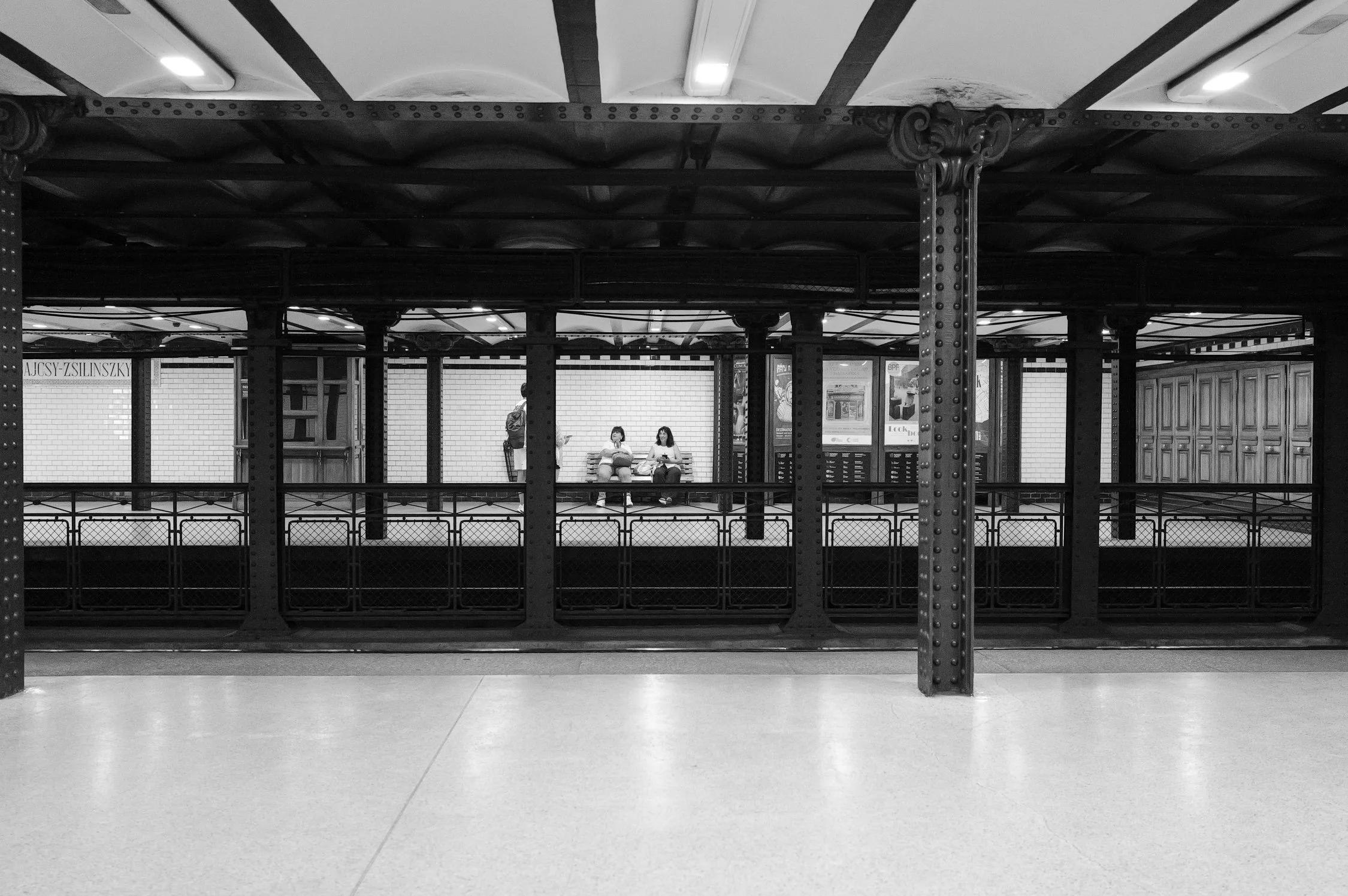 Deux personnes assises sur un banc à une station de métro, vues à travers une barrière en métal, sous une plateforme de métro.