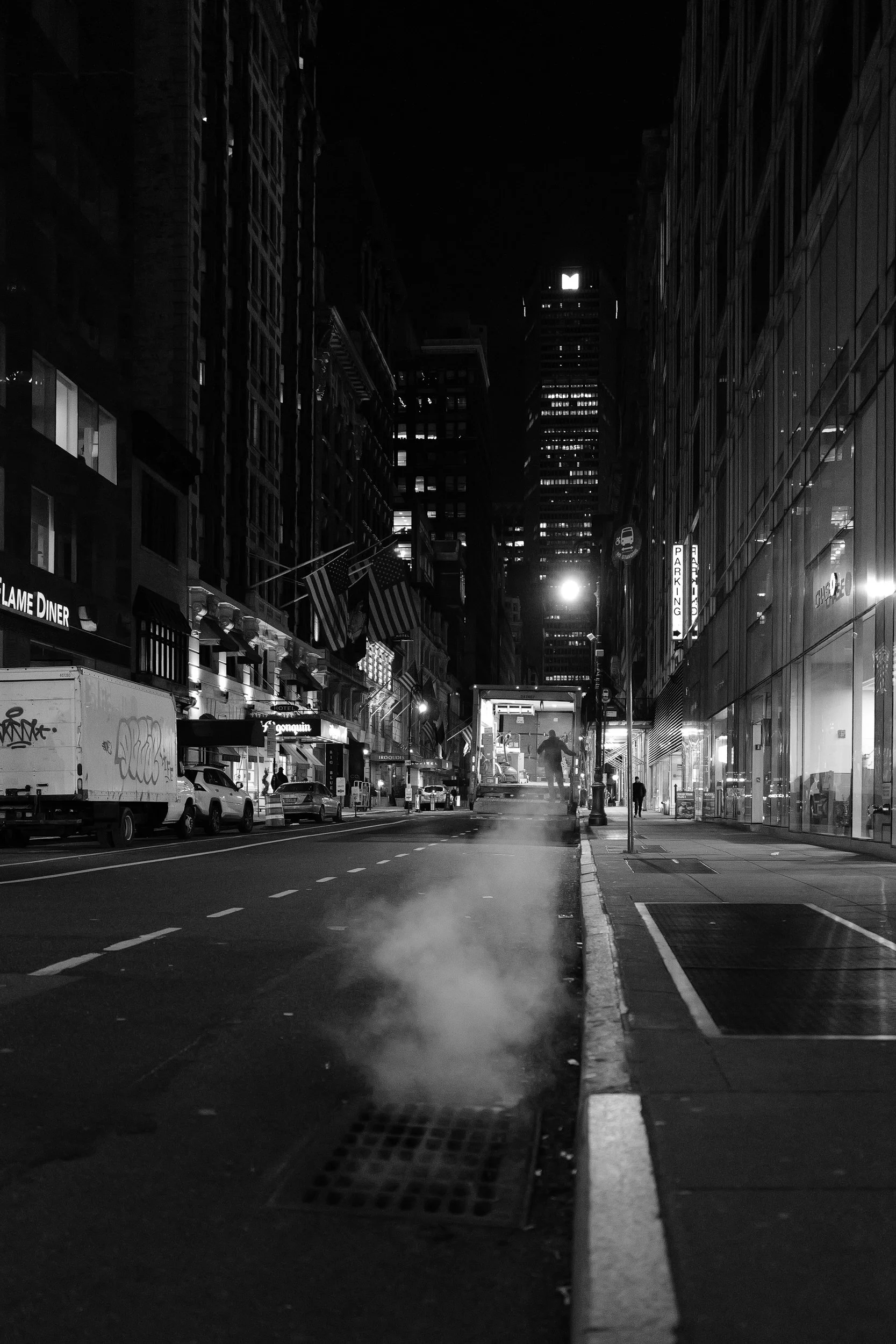 Vue nocturne d'une rue urbaine avec des bâtiments illuminés, un véhicule de nettoyage ou de construction émettant de la vapeur, et des enseignes lumineuses, en noir et blanc.