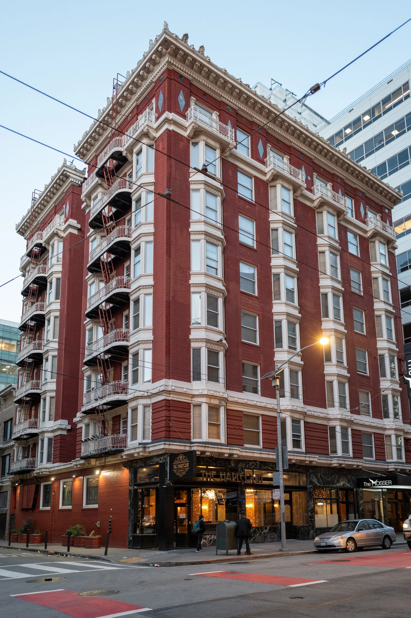 Bâtiment de style victorien en brique rouge avec des balcons en fer forgé, situé dans un environnement urbain, avec des lampadaires allumés et quelques personnes devant le bâtiment.