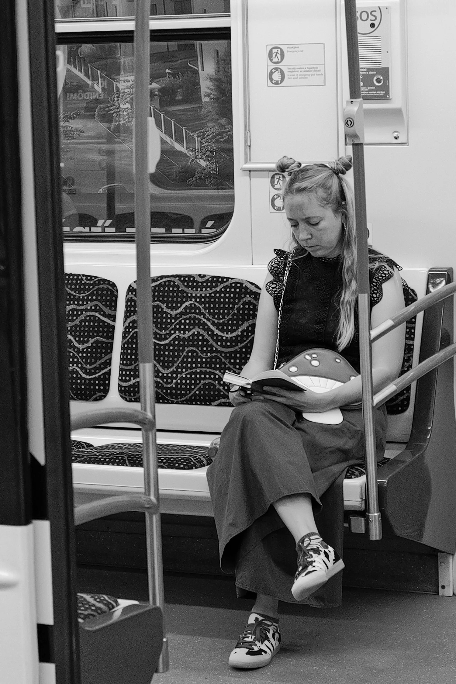 Une jeune fille aux cheveux blonds attachés en deux chignons, assise dans un métro, lit un livre, portant une robe noire avec des baskets à motifs.