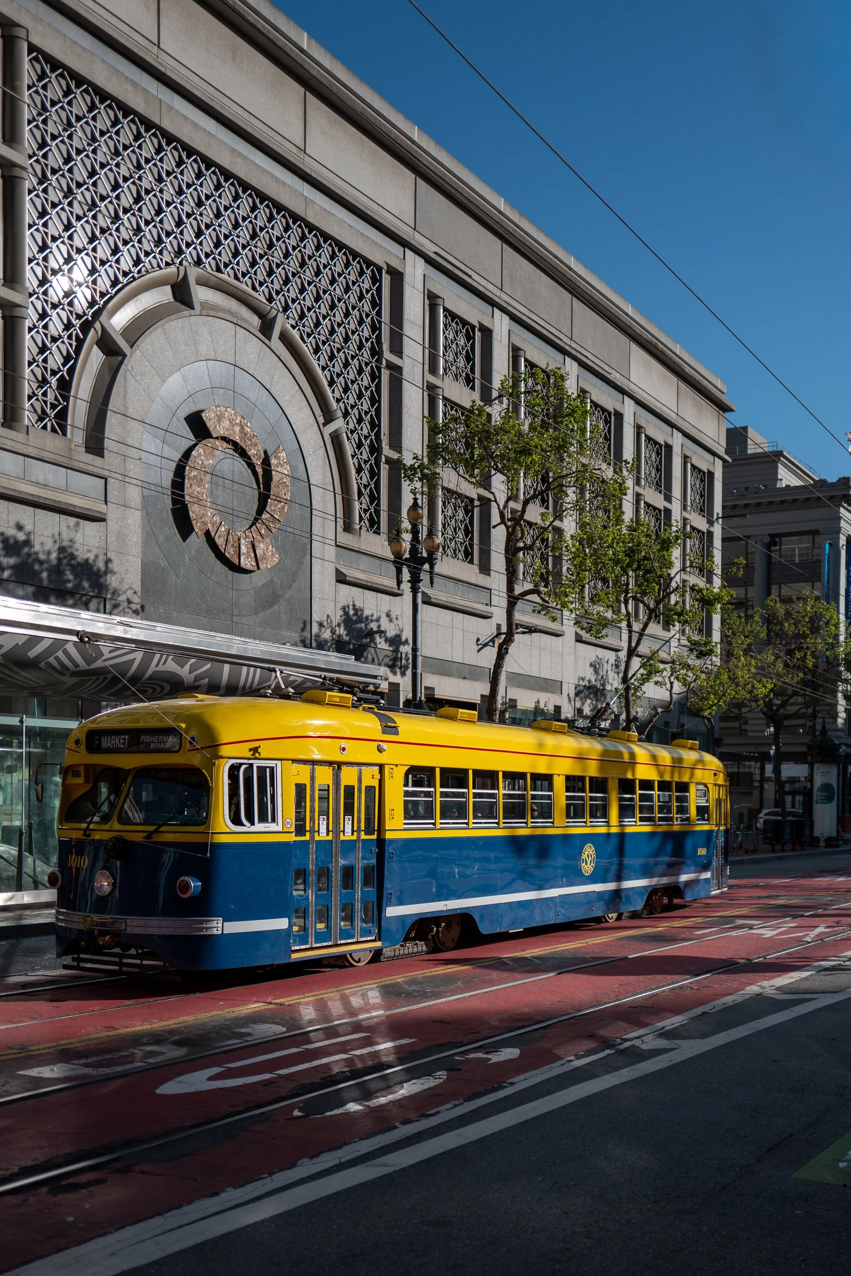 Un tramway jaune et bleu stationne sur une voie en ville avec un bâtiment moderne en arrière-plan, arbres et lignes électriques au-dessus.