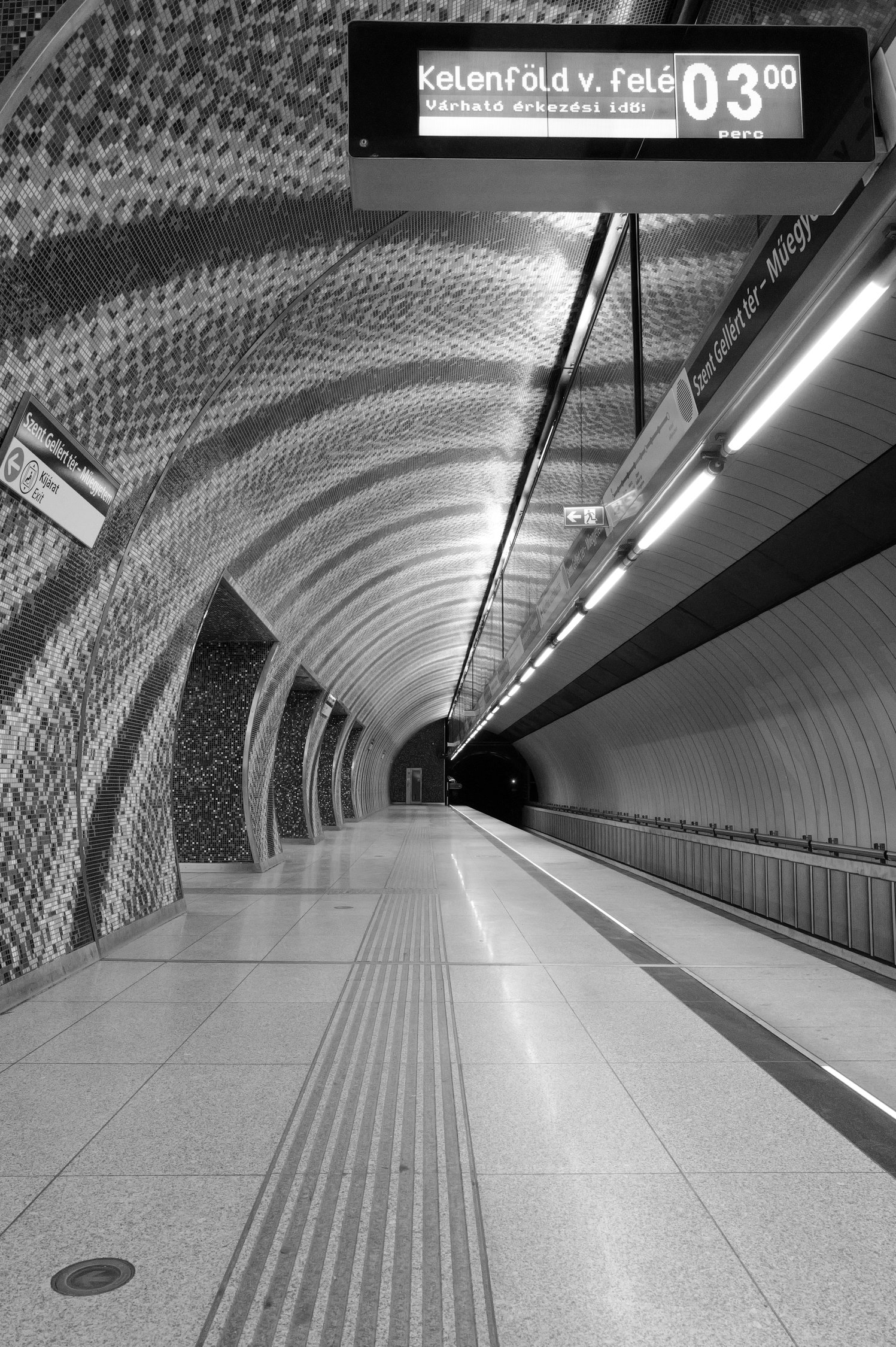 Une station de métro vide avec un tunnel courbé en mosaïque, des signes d'orientation, un panneau d'affichage avec des informations en hongrois, et un sol en carreaux clairs.