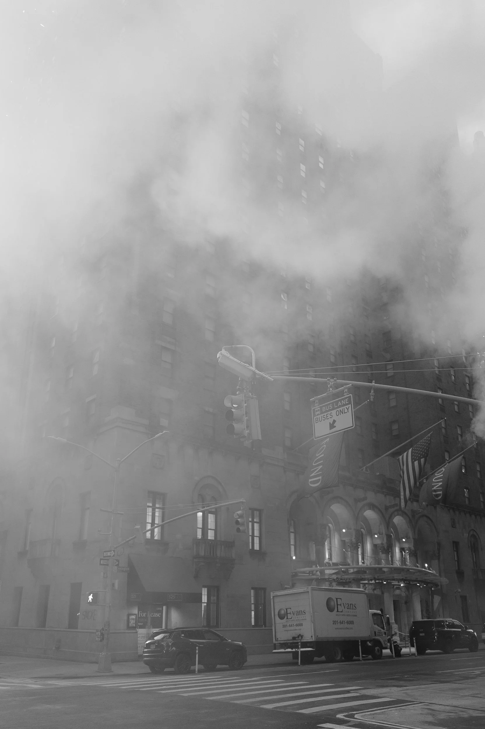Une rue en ville avec un bâtiment ancien, un camion de déménagement, des voitures, des drapeaux et de la vapeur s'élevant dans l'air.
