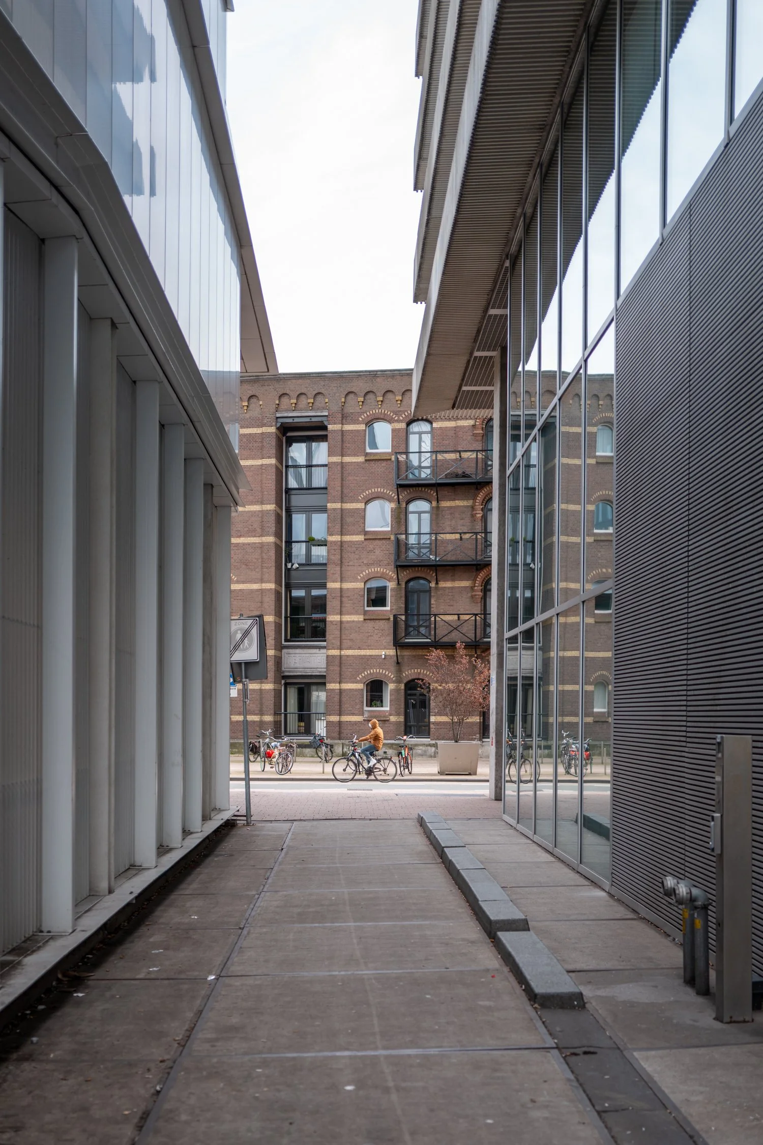 vue d'une rue urbaine avec un bâtiment en briques, des vélos, et une personne à vélo, prise depuis un passage étroit entre deux bâtiments modernes.