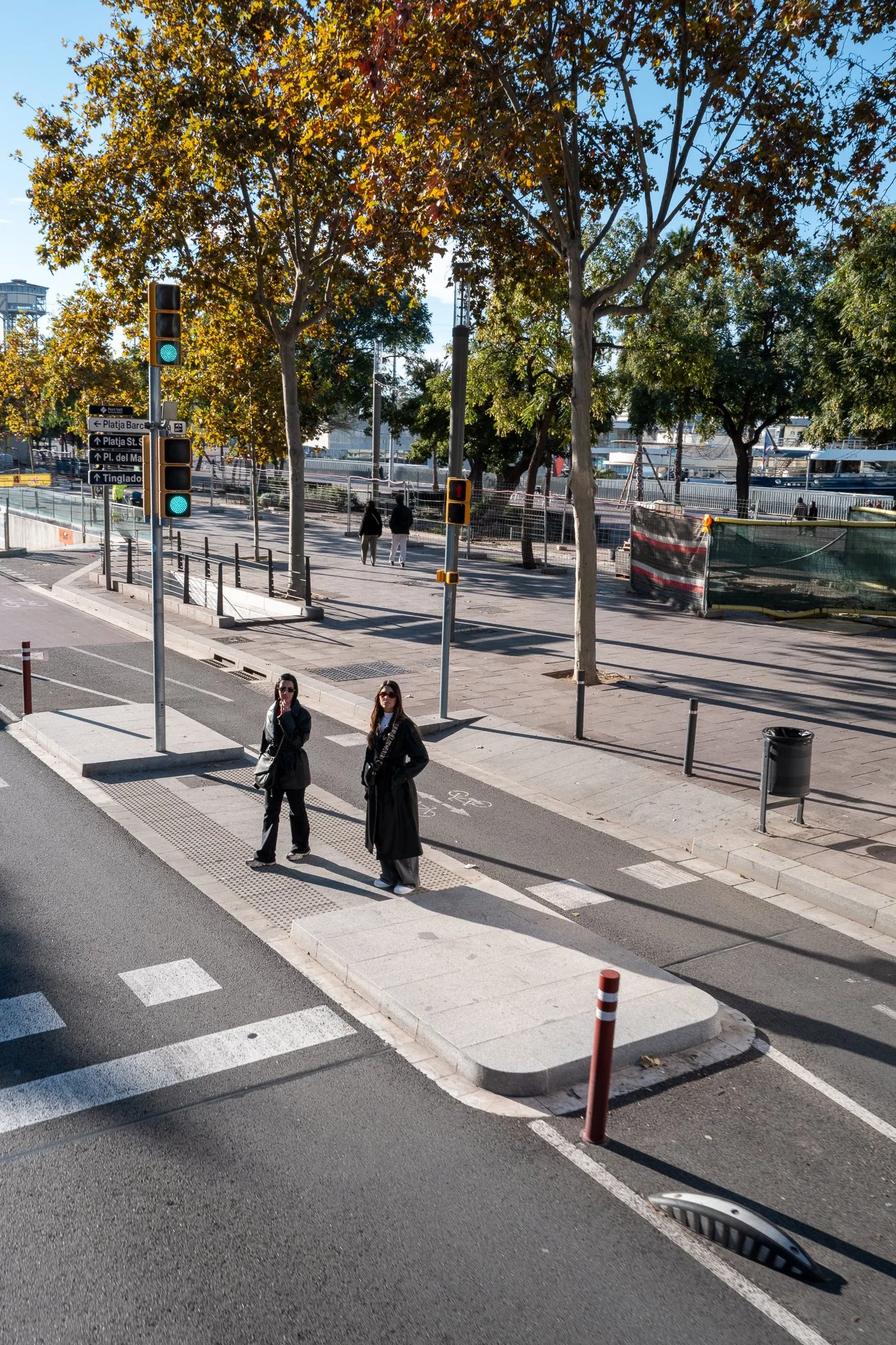 Deux femmes attendent au passage pour piétons face à un feu de circulation vert, dans une rue urbaine bordée d'arbres à feuillage automnal. Le ciel est clair, et le décor urbain inclut des panneaux de signalisation, un caniveau et une poubelle.