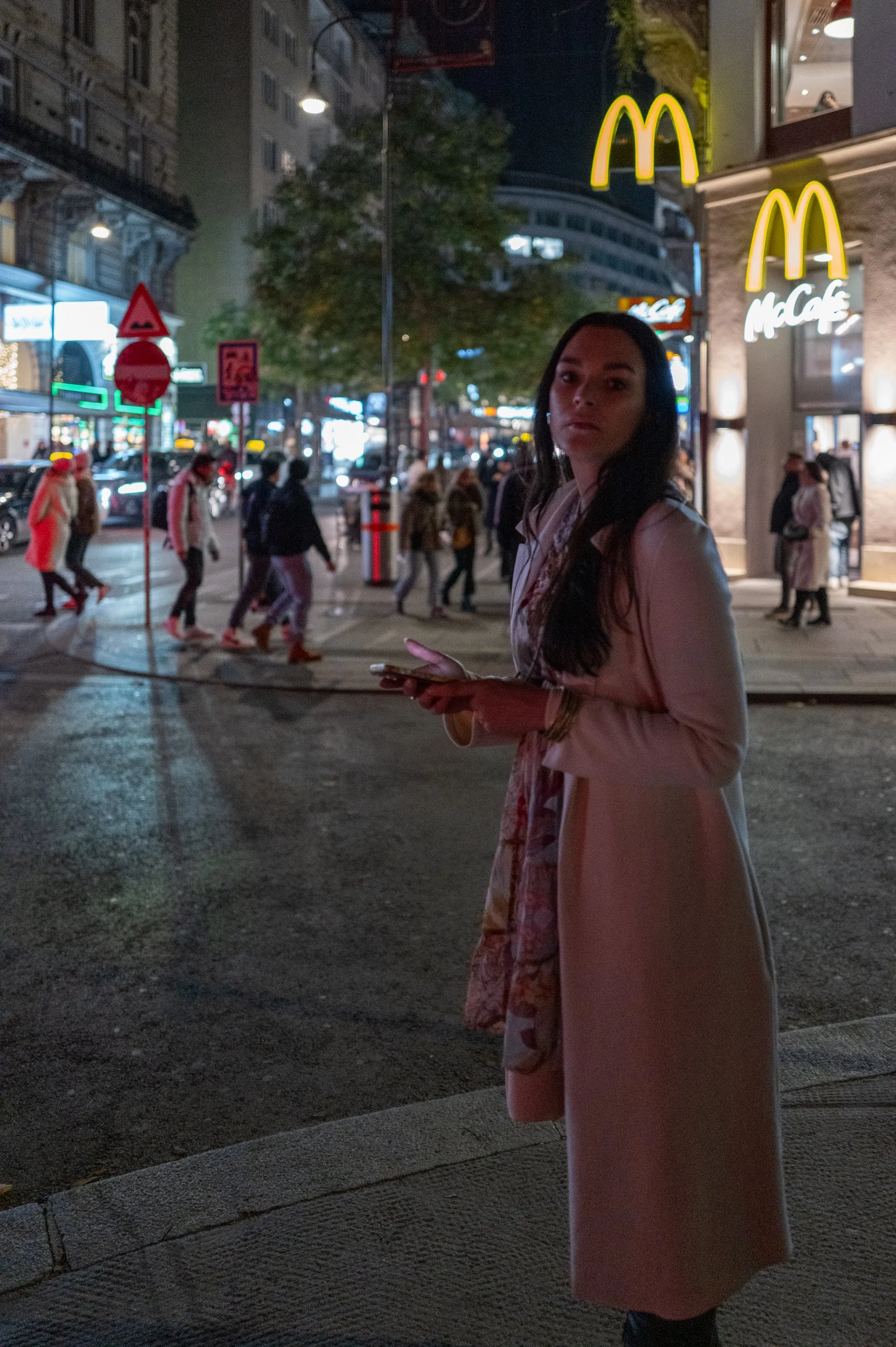 Une femme avec un long manteau rose foncé se tient sur le trottoir devant un McDonald's la nuit. En arrière-plan, il y a des piétons traversant la rue et des enseignes lumineuses dans une ville animée.
