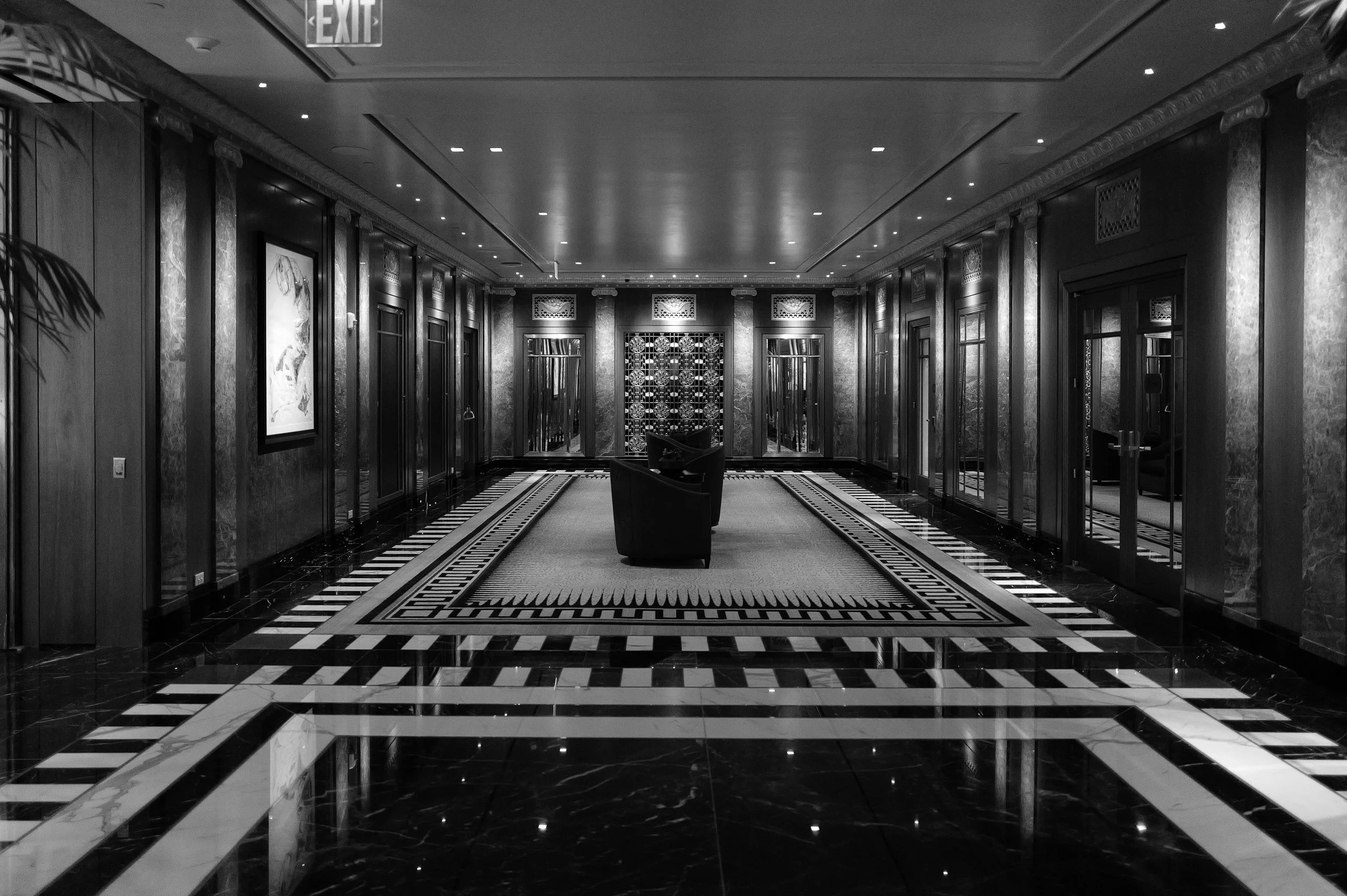 Intérieur d'un hall d'hôtel ou de bâtiment élégant avec des murs en marbre, un tapis central, des fauteuils, et des décorations sophistiquées, en noir et blanc.