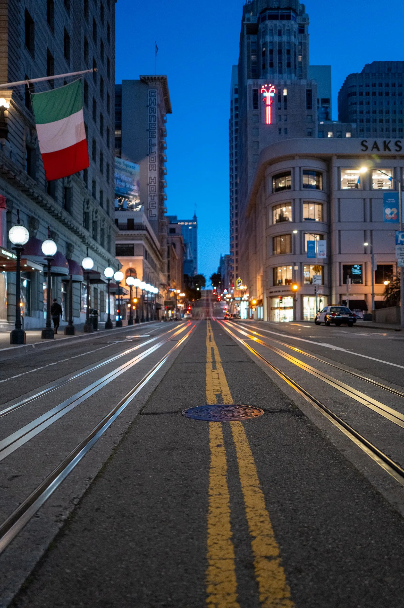 Une rue urbaine en ville avec des bâtiments illuminés, des lampadaires, une bannière de drapeau italien, et des rails pour tramway, prise au crépuscule.