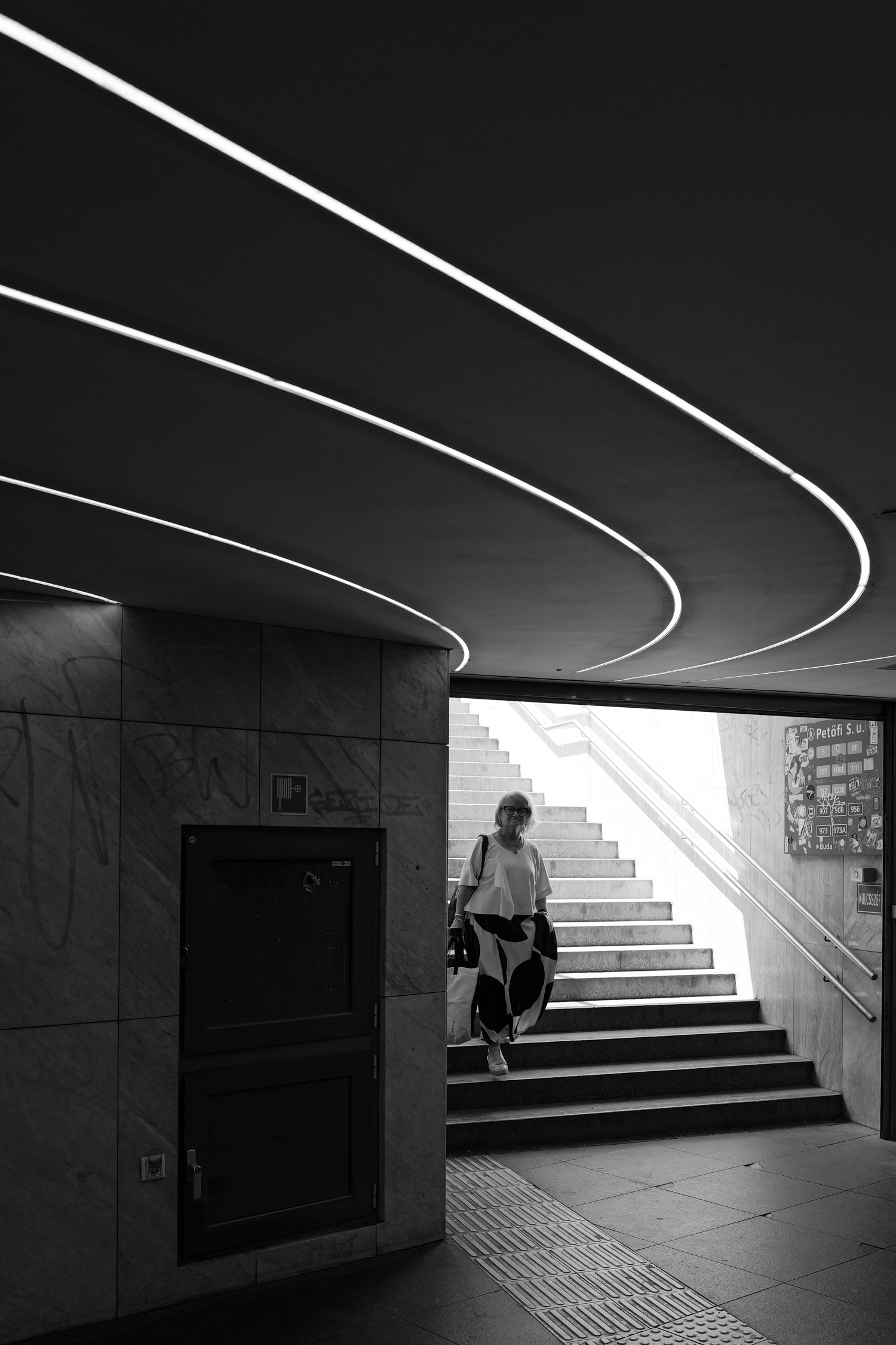 Une personne portant un vêtement à motifs noirs et blancs descend un escalier dans une station de métro ou une gare. La scène est en noir et blanc, avec des lumières modernes au plafond.