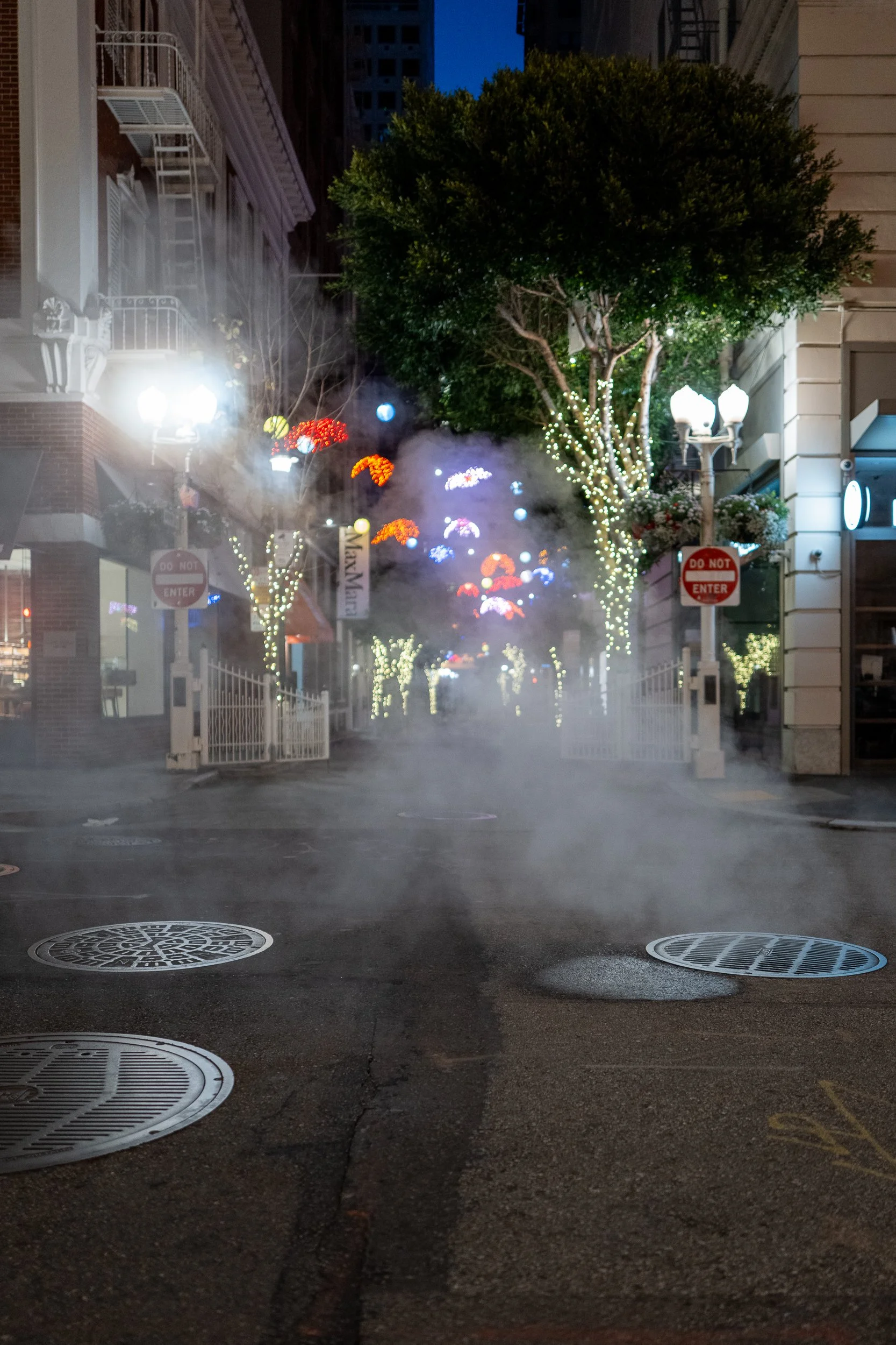 Une rue urbaine la nuit, décorée avec des lumières de Noël, des arbres illuminés et de la brume ou vapeur sur le sol. Il y a des panneaux 'Interdiction d'entrer' et des lampadaires.