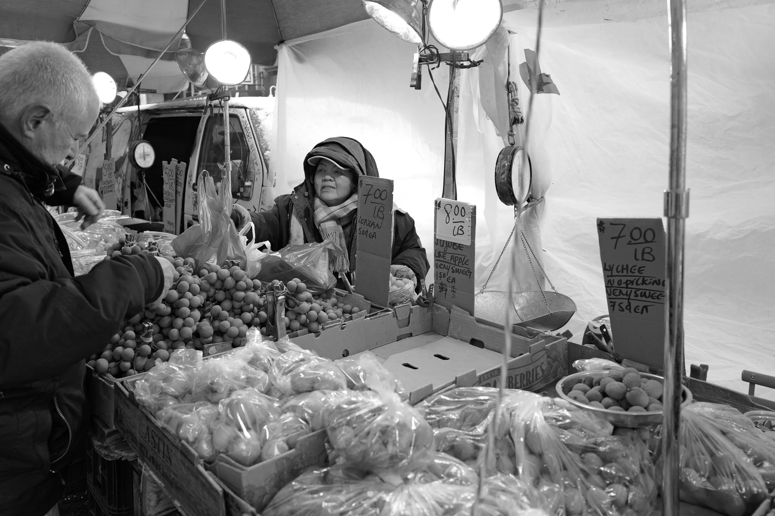 Vendeur vendant des fruits, notamment des cerises, à un client dans un marché en plein air, avec des signaux de prix affichés en euros.