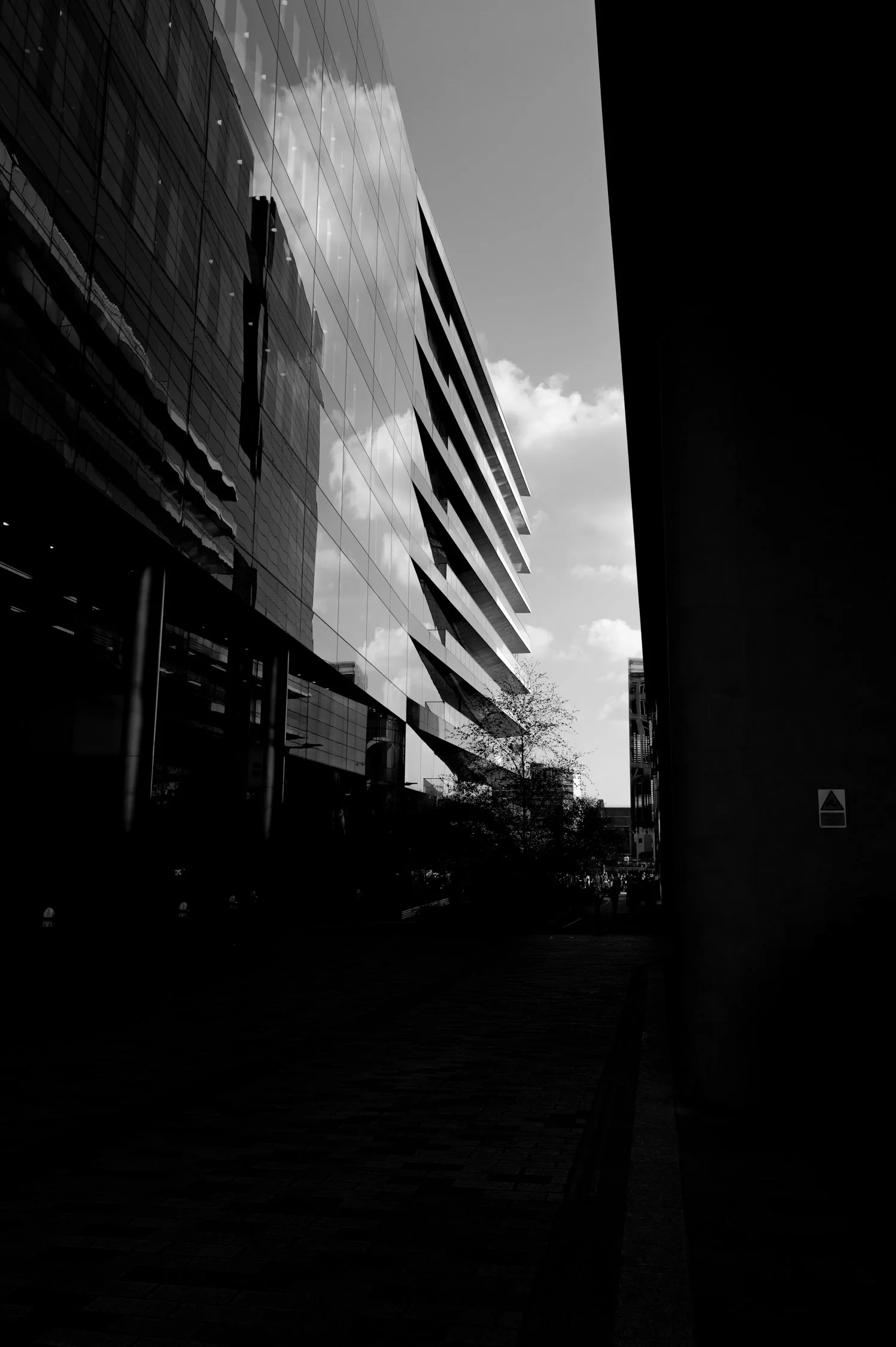 Photo en noir et blanc d'un bâtiment moderne avec une façade en verre, une rue pavée et le ciel avec quelques nuages.