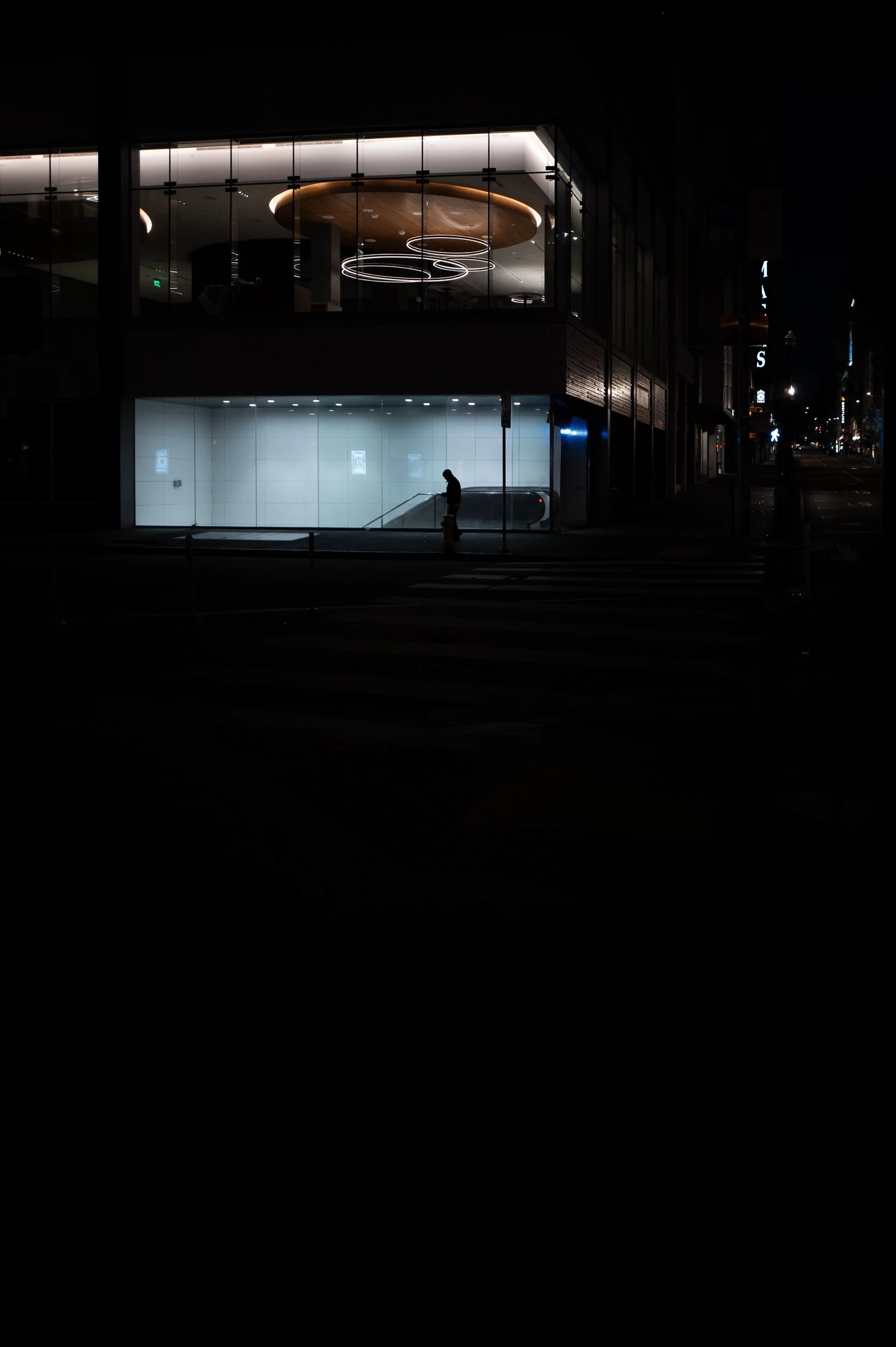 Une façade de bâtiment moderne avec deux niveaux éclairés, une personne marche sur un trottoir dans une rue nocturne