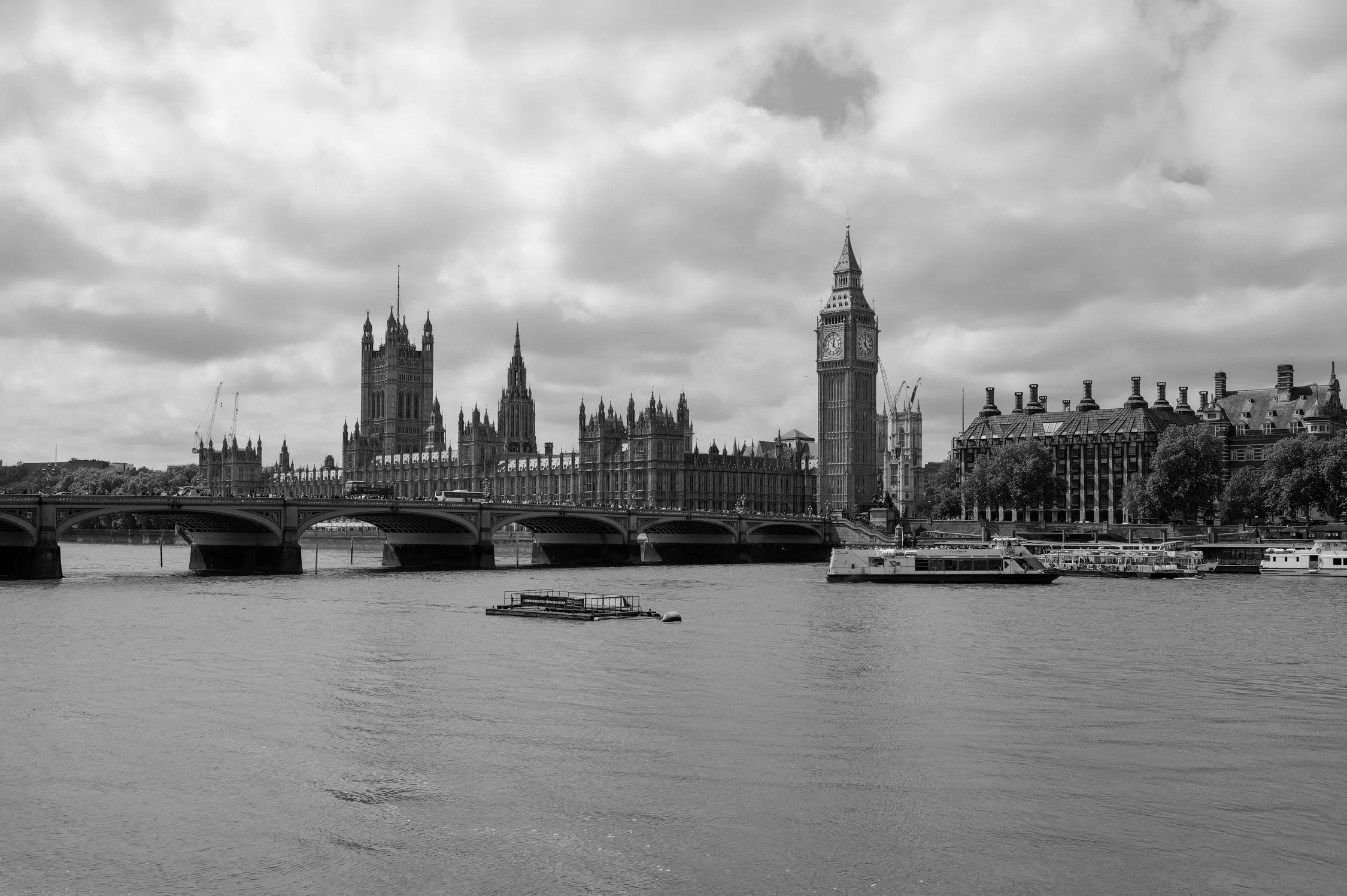Vue d'un panorama de Londres en noir et blanc, avec le fleuve autour et la silhouette du Big Ben et du palais de Westminster en arrière-plan, sous un ciel nuageux.