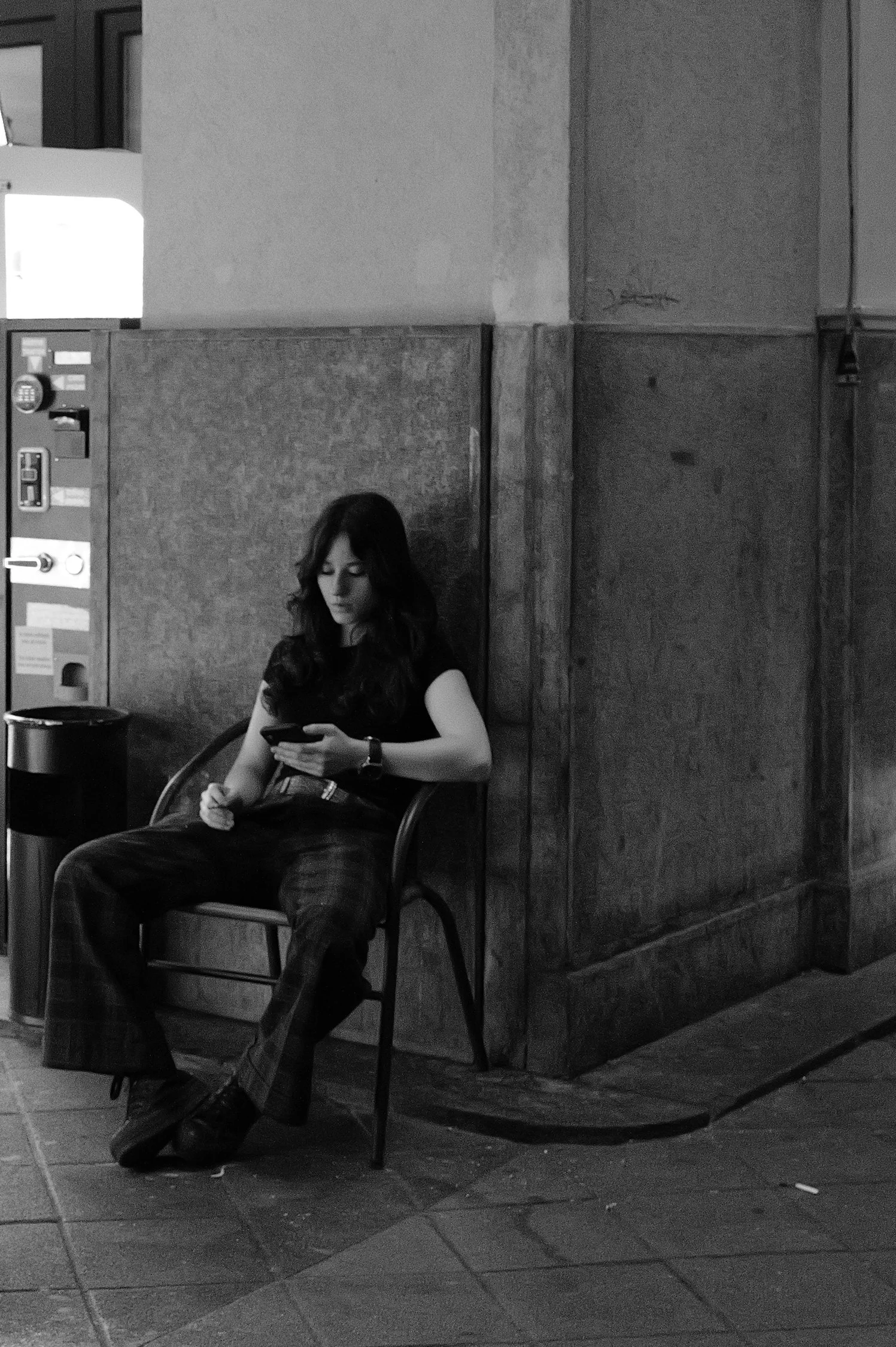 Une femme assise sur une chaise dans un endroit intérieur, regardant son téléphone portable. Elle porte une tenue noire avec un bracelet à la main gauche, ses cheveux sont longs et ondulés, et elle est devant un mur en bois.