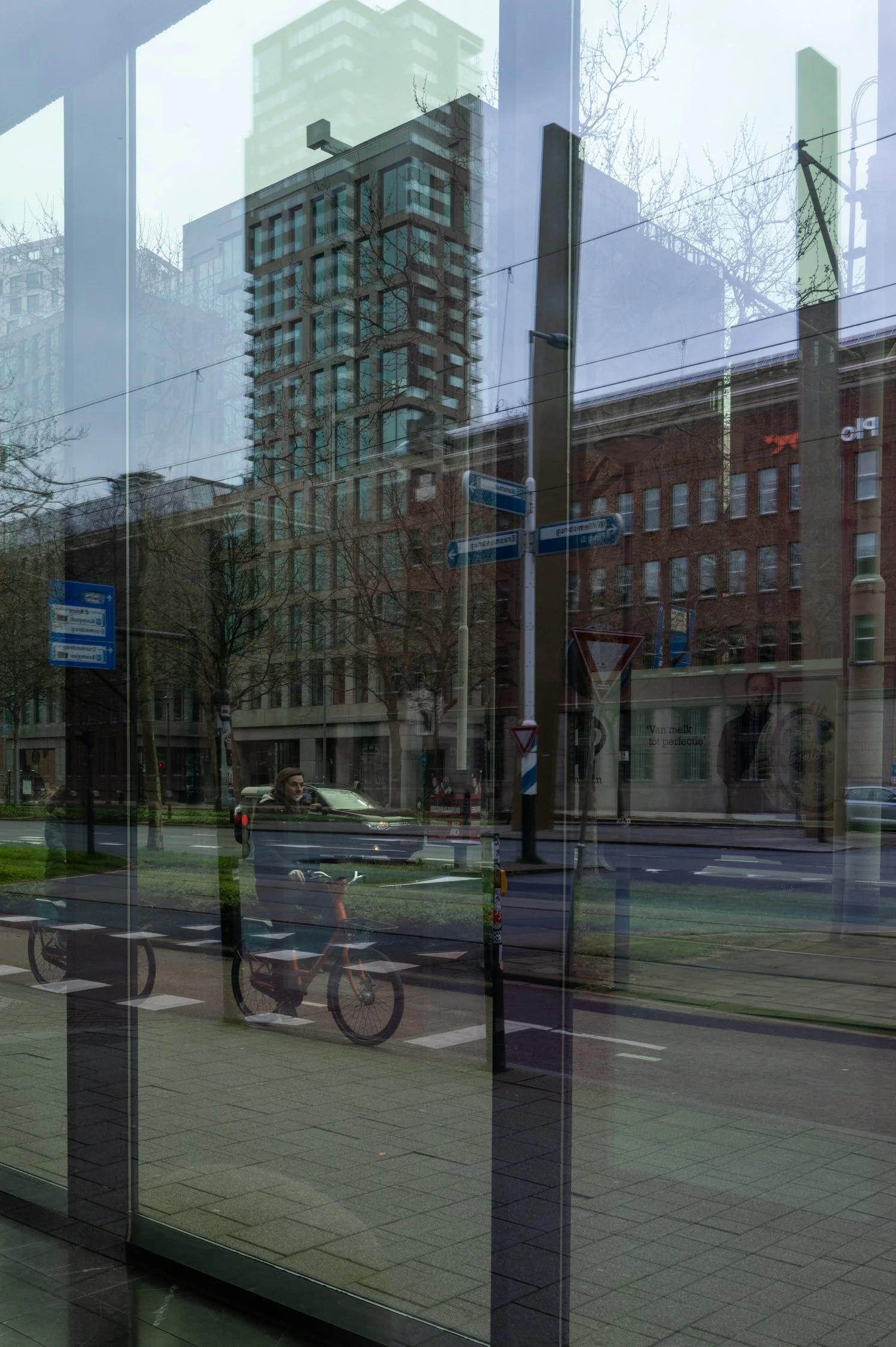 Reflet d’un bâtiment moderne et d’un arbre sans feuilles dans une vitrine en verre, avec un trottoir, une bicyclette, des panneaux de signalisation, et des bâtiments en arrière-plan.