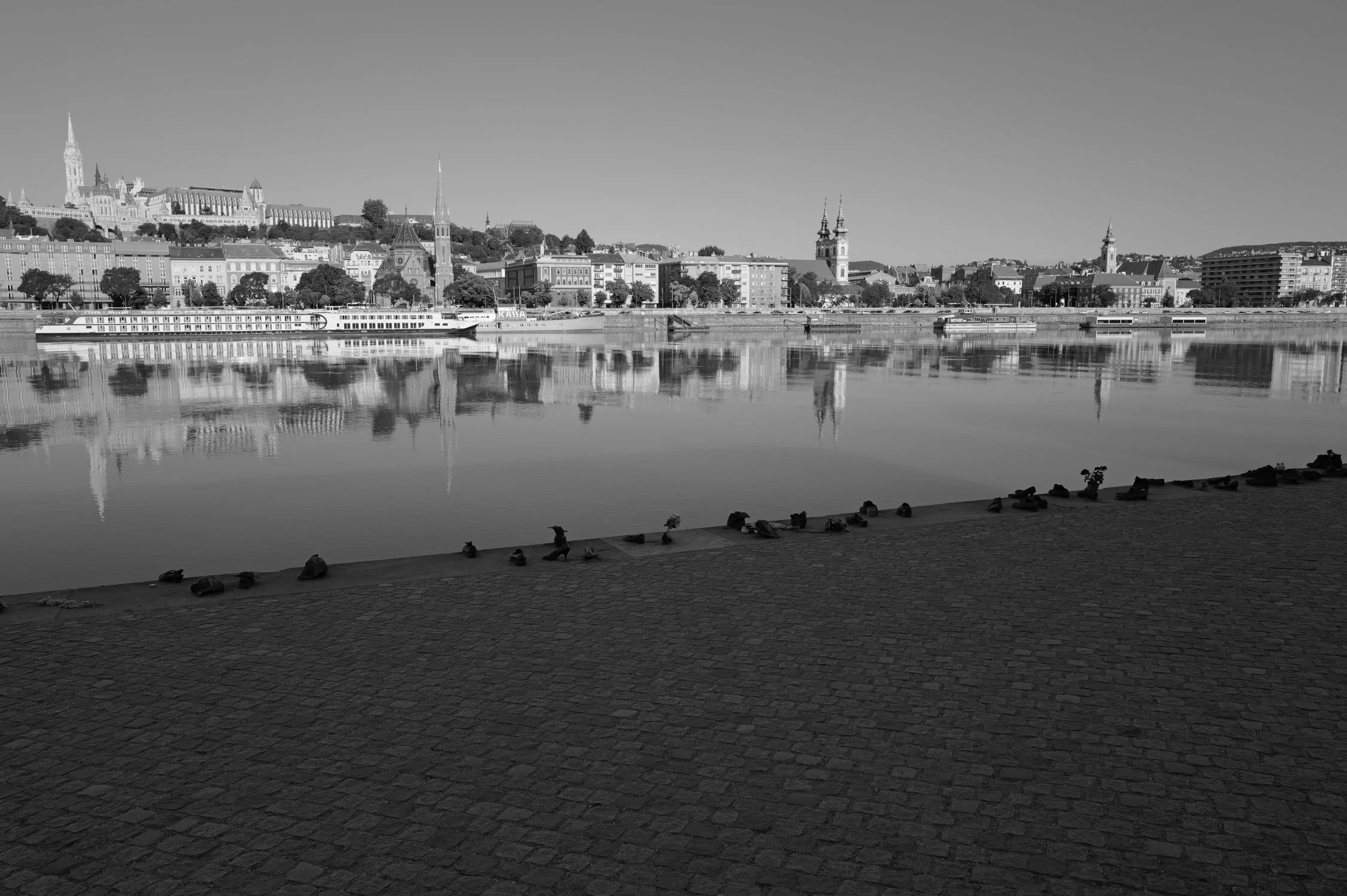 Vue panoramique d'une ville avec des bâtiments historiques, un fleuve en premier plan, plusieurs bateaux, et des e charbon à l'avant-plan. Image en noir et blanc.