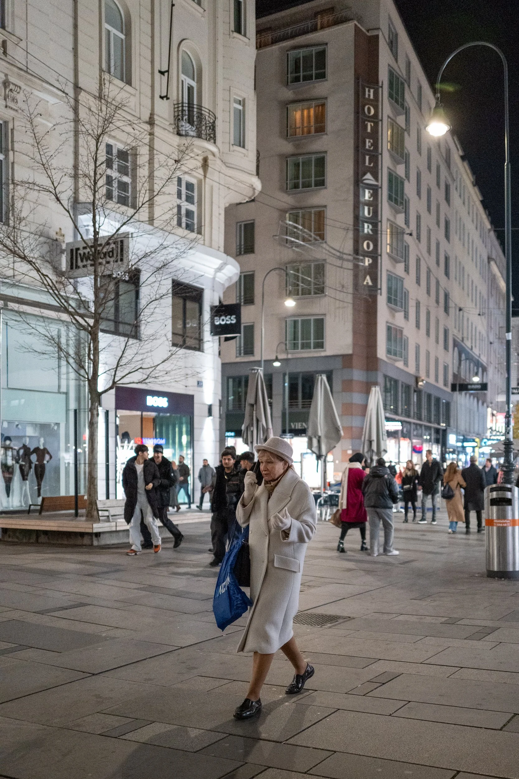 Une femme habillée en beige marche dans une rue pavée de nuit, entourée de passants, avec des magasins et hôtels en arrière-plan, éclairés par des lampadaires.