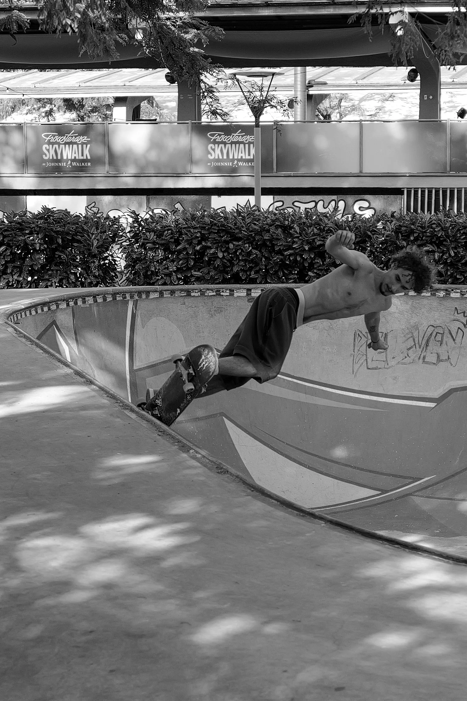 Jeune homme faisant du skate dans un skatepark, souriant et portant un pantalon, sans chemise, avec des graffitis sur la paroi du skatepark.