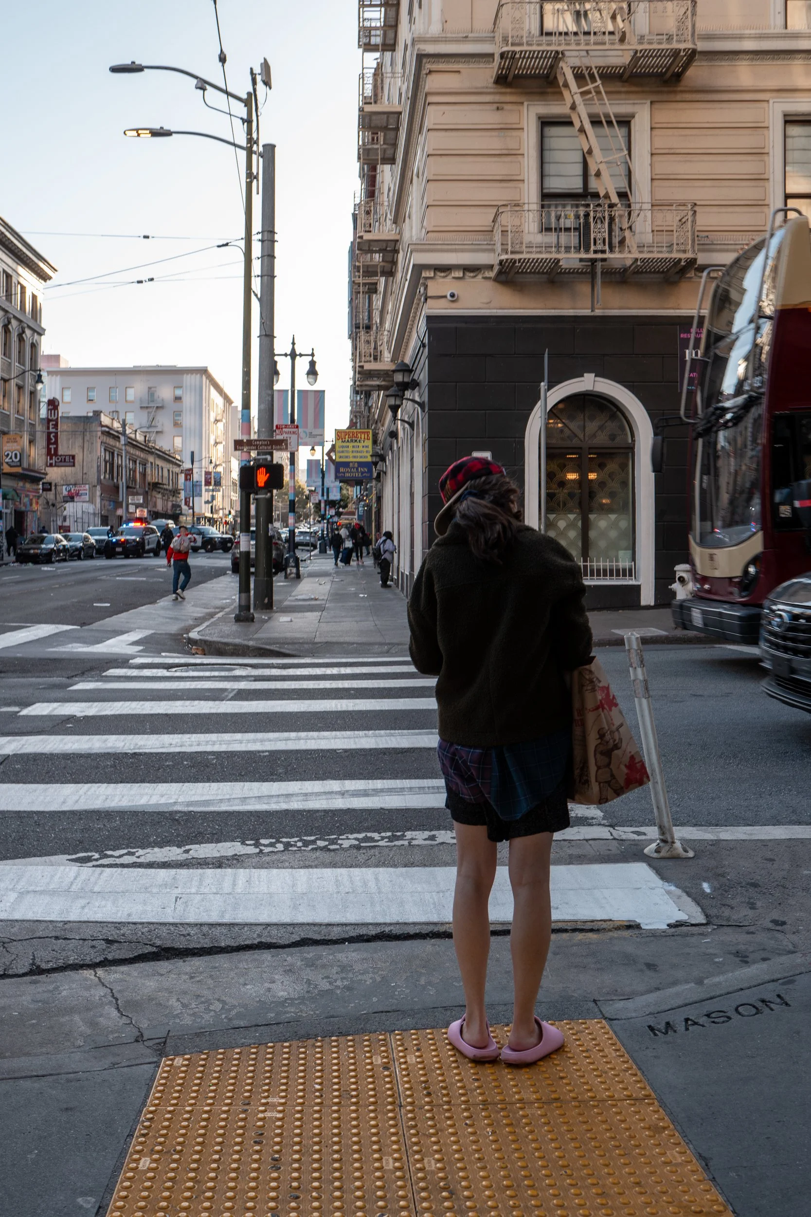 Une jeune femme en vêtements décontractés et maillot de bain, portant une casquette, se tient à un passage piéton au coin d'une rue urbaine. Elle porte des pantoufles roses et tient un sac en papier sur le bras.