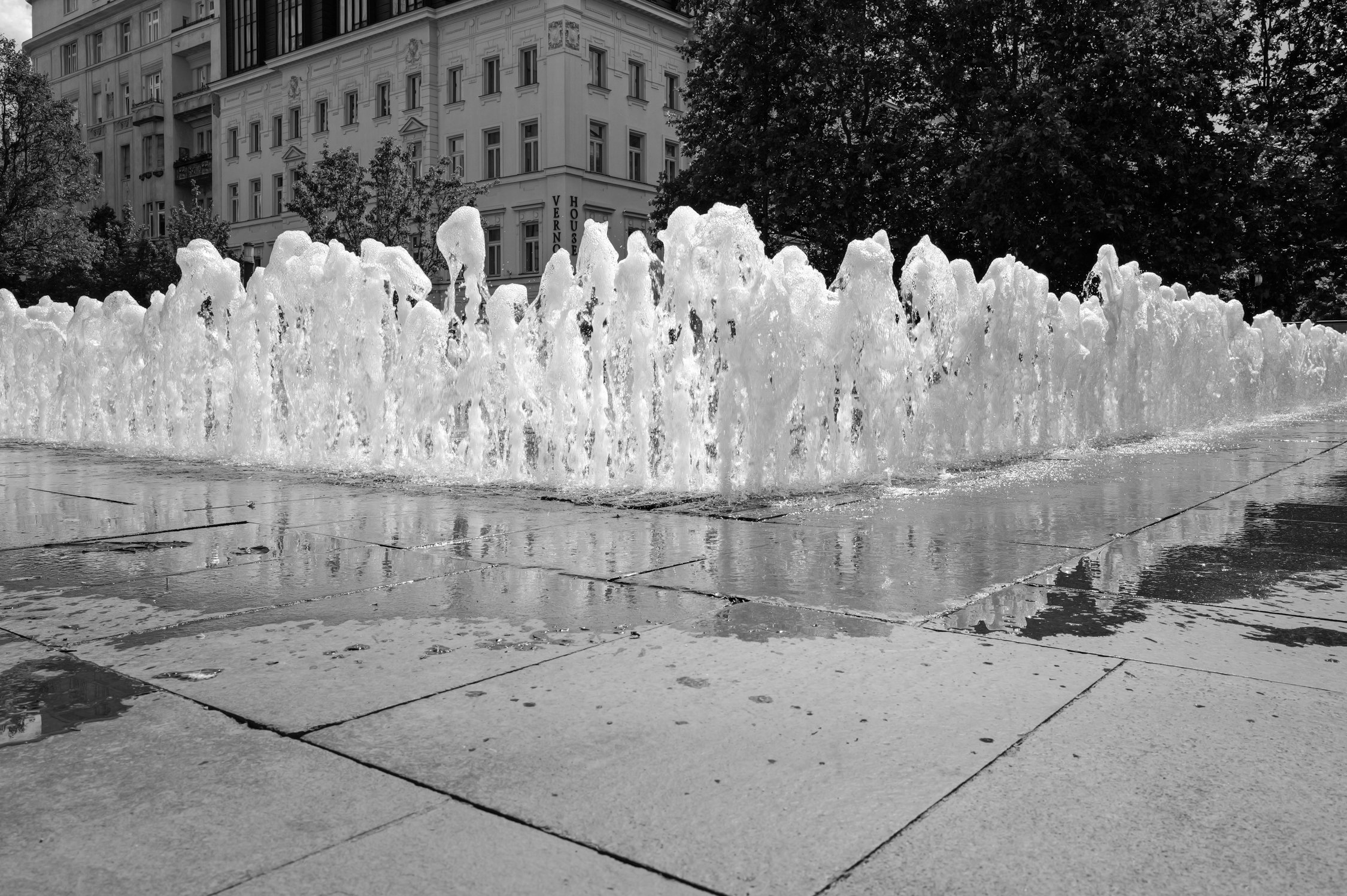 Une fontaine avec de l'eau jaillissant, des bâtiments résidentiels en arrière-plan et des arbres environnants, vue en noir et blanc.