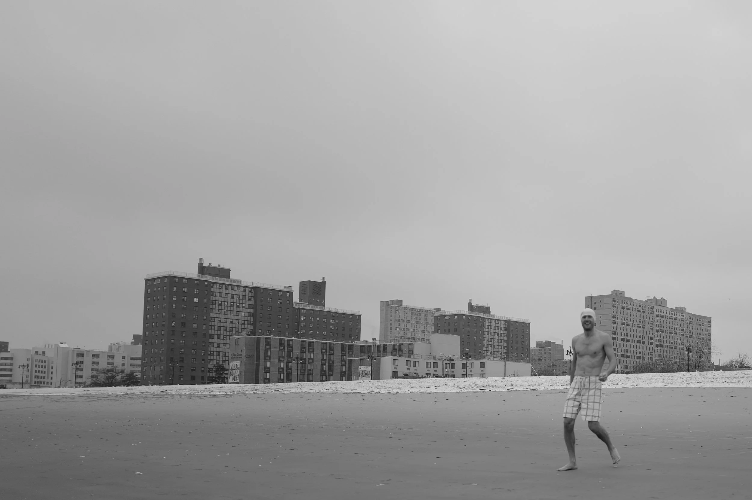 Un homme souriant portant un bonnet, sans chemise et en short à carreaux, marche sur la plage sous un ciel nuageux avec des immeubles en arrière-plan.