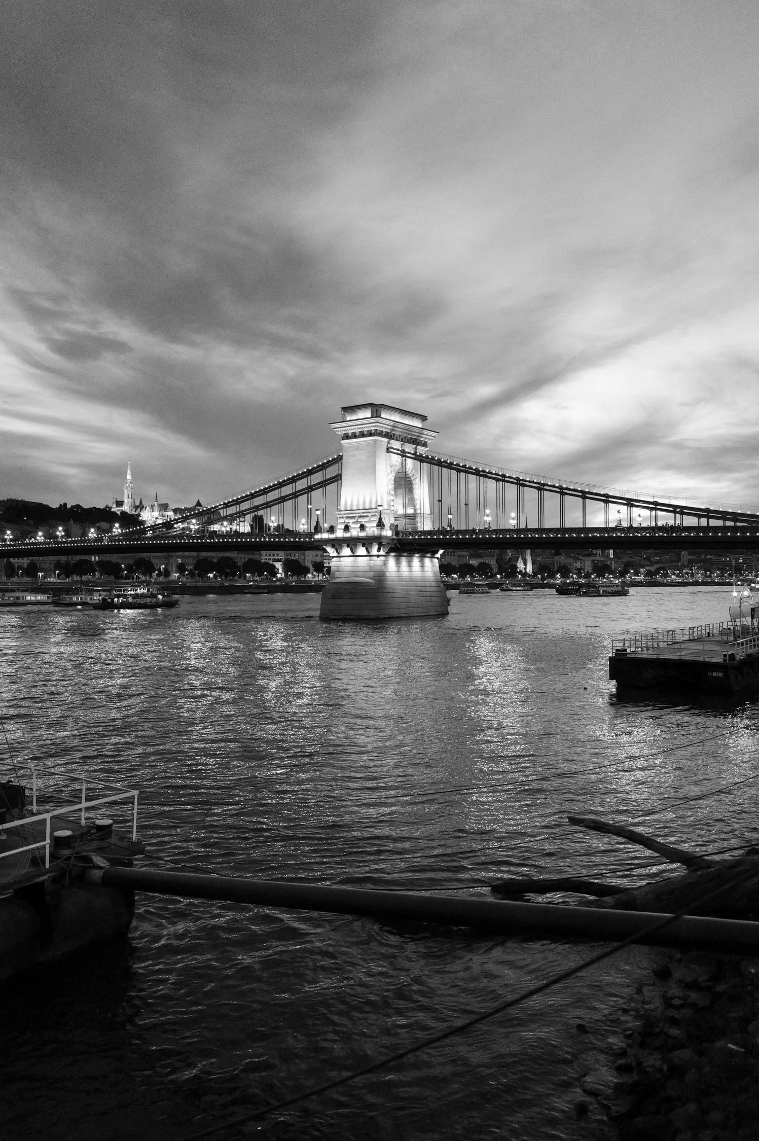 Vue du pont de Budapest la nuit, avec le Danube en premier plan et des bâtiments illuminés en arrière-plan, sous un ciel nuageux.