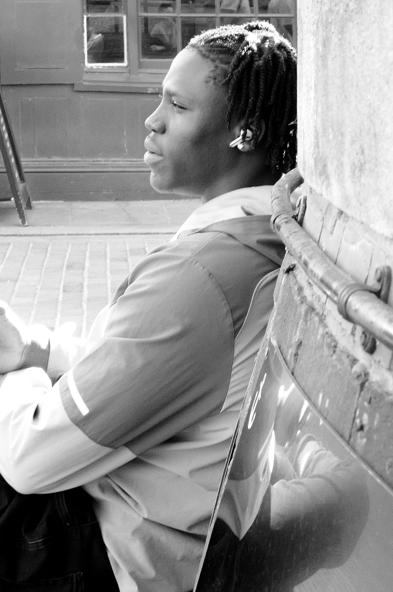 Un jeune homme assis, vu en profil gauche, portant un blazer et une chemise, avec des dreadlocks, assis contre un mur en pierre, dans un environnement intérieur ou extérieur.