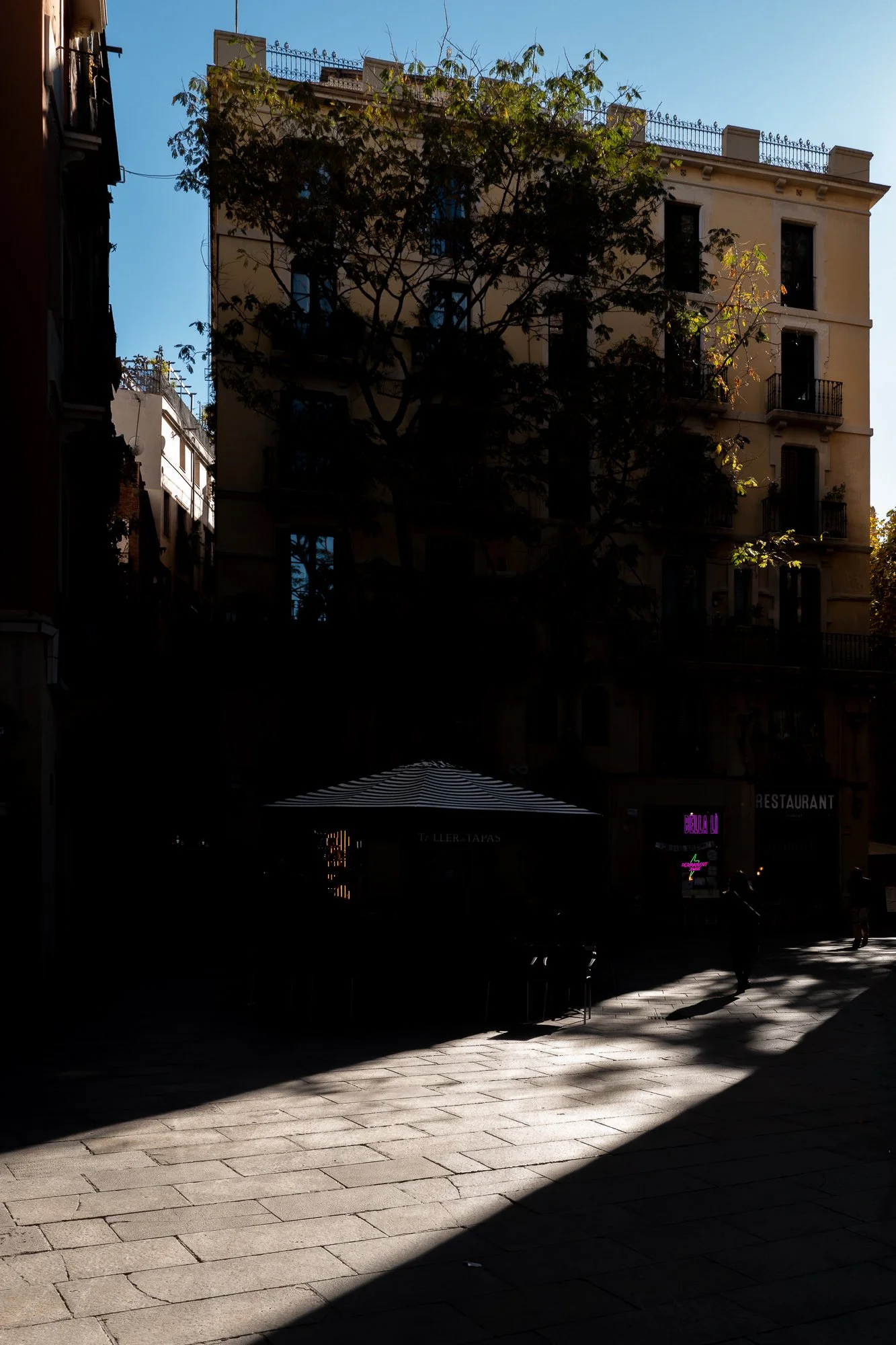 Une scène urbaine au coucher du soleil avec un bâtiment de plusieurs étages, des arbres, une terrasse de café avec une pergola rayée, et quelques personnes marchant sur le trottoir. L'éclairage crée des ombres longues.
