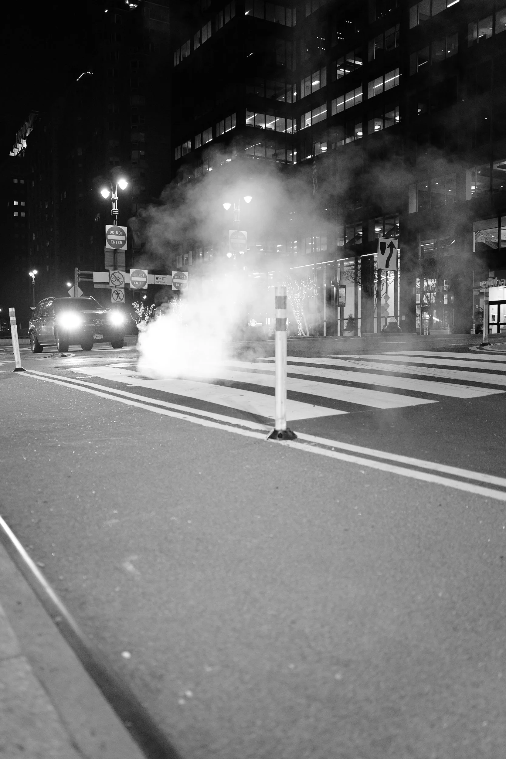 Une rue urbaine la nuit avec un fort éclairage, des voitures à l'arrêt, des panneaux designalisation, un passage piéton et de la vapeur ou de la fumée s'échappant du sol.
