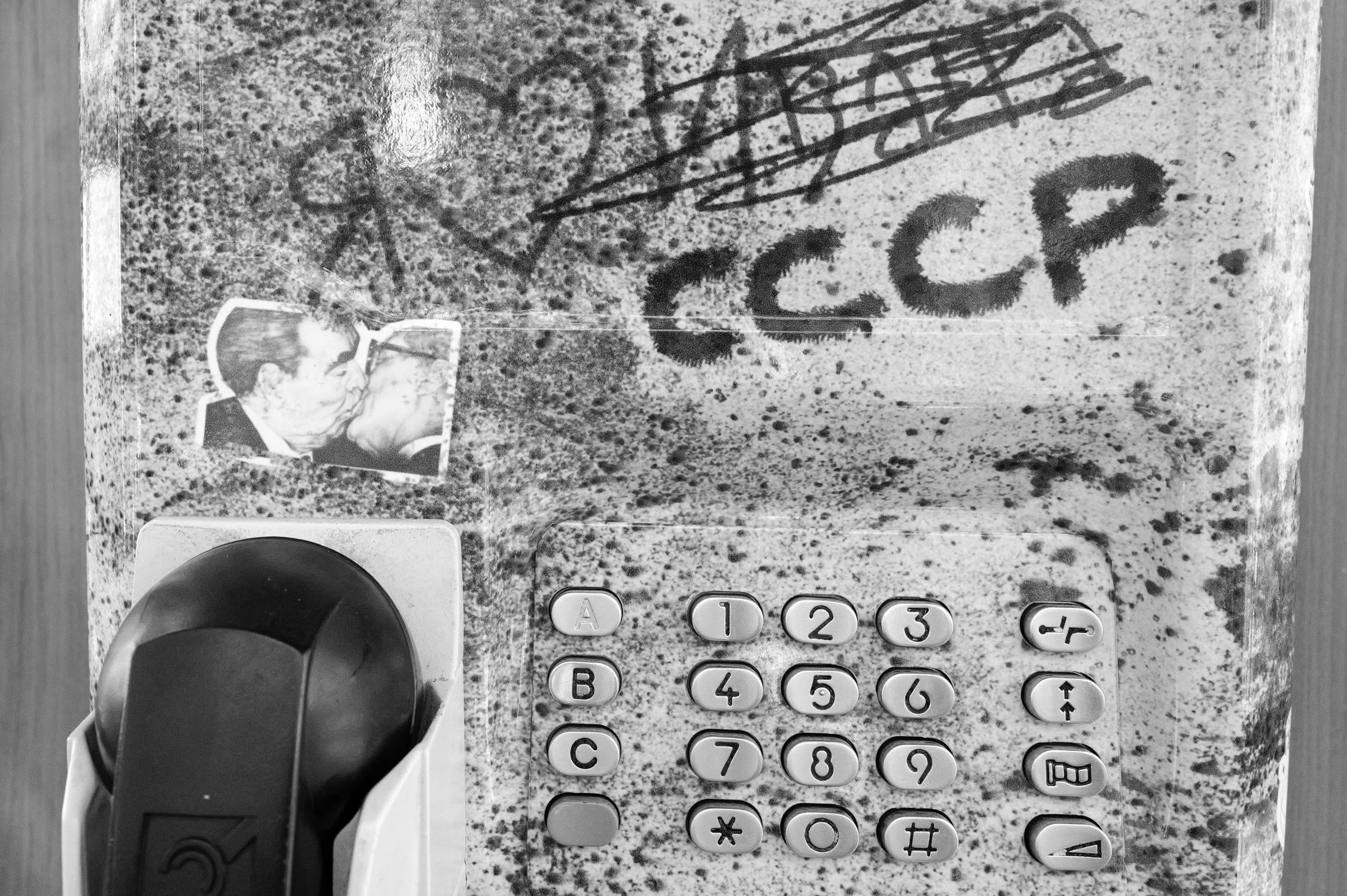 Un téléphone fixe avec un clavier numérique, un micro à gauche, une photo de deux personnes qui s'embrassent collée à l'écran, et des inscriptions manuscrites telles que "NO" et d'autres dessins au-dessus du clavier. Le tout est posé sur une surface 