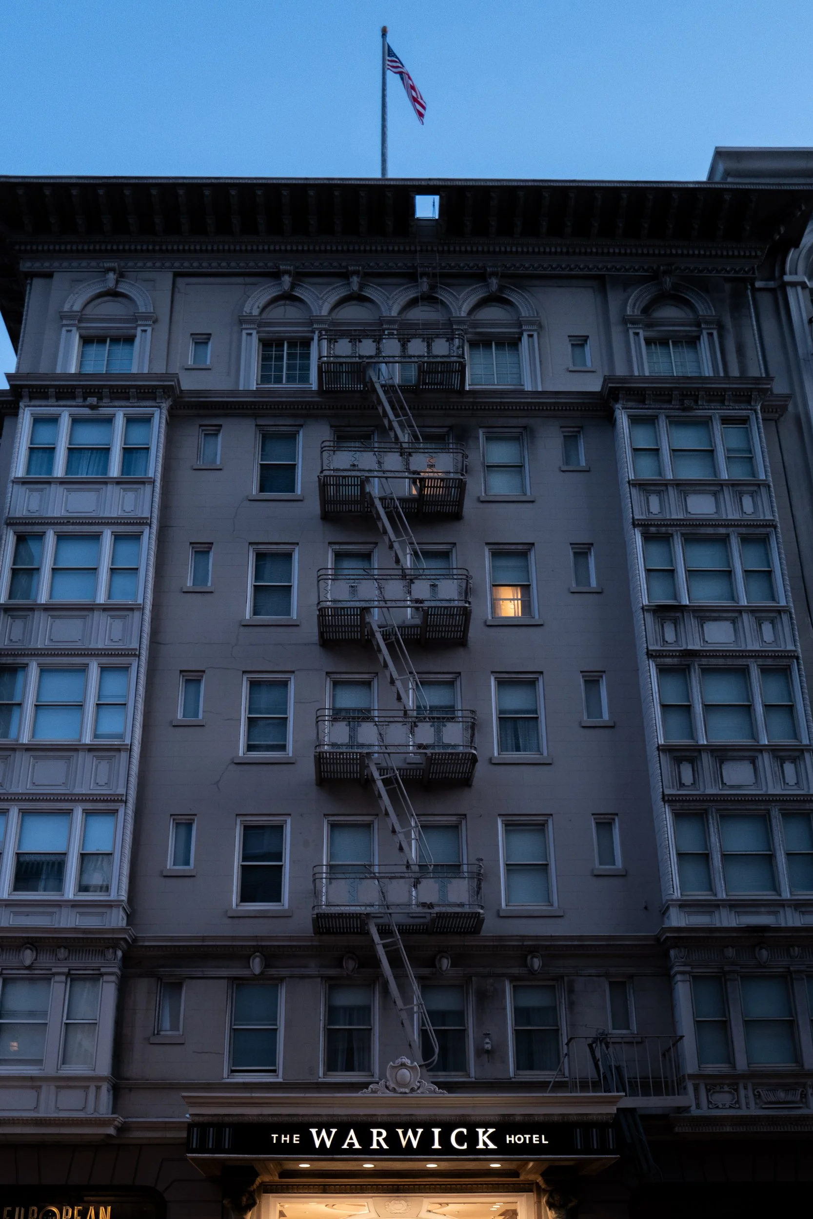 Un bâtiment en pierre avec un drapeau américain en haut, plusieurs fenêtres et balcons, et une enseigne lumineuse indiquant "The Warwick Hotel" à la base.