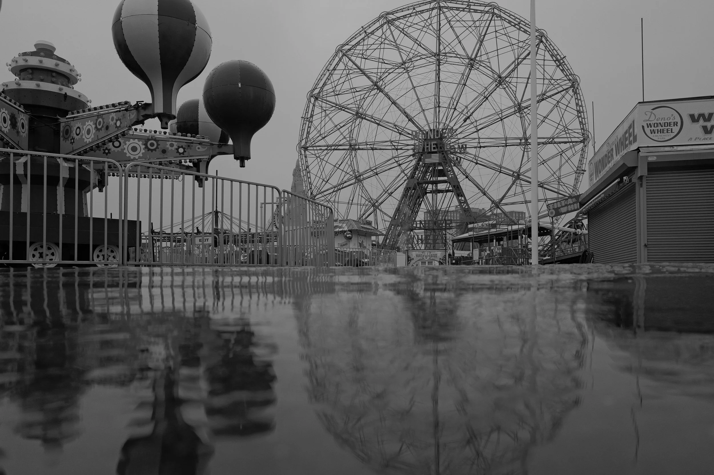 Une fête foraine avec des manèges et une grande roue, vue depuis une prise de vue au niveau du sol avec une flaque d'eau qui reflète les structures.