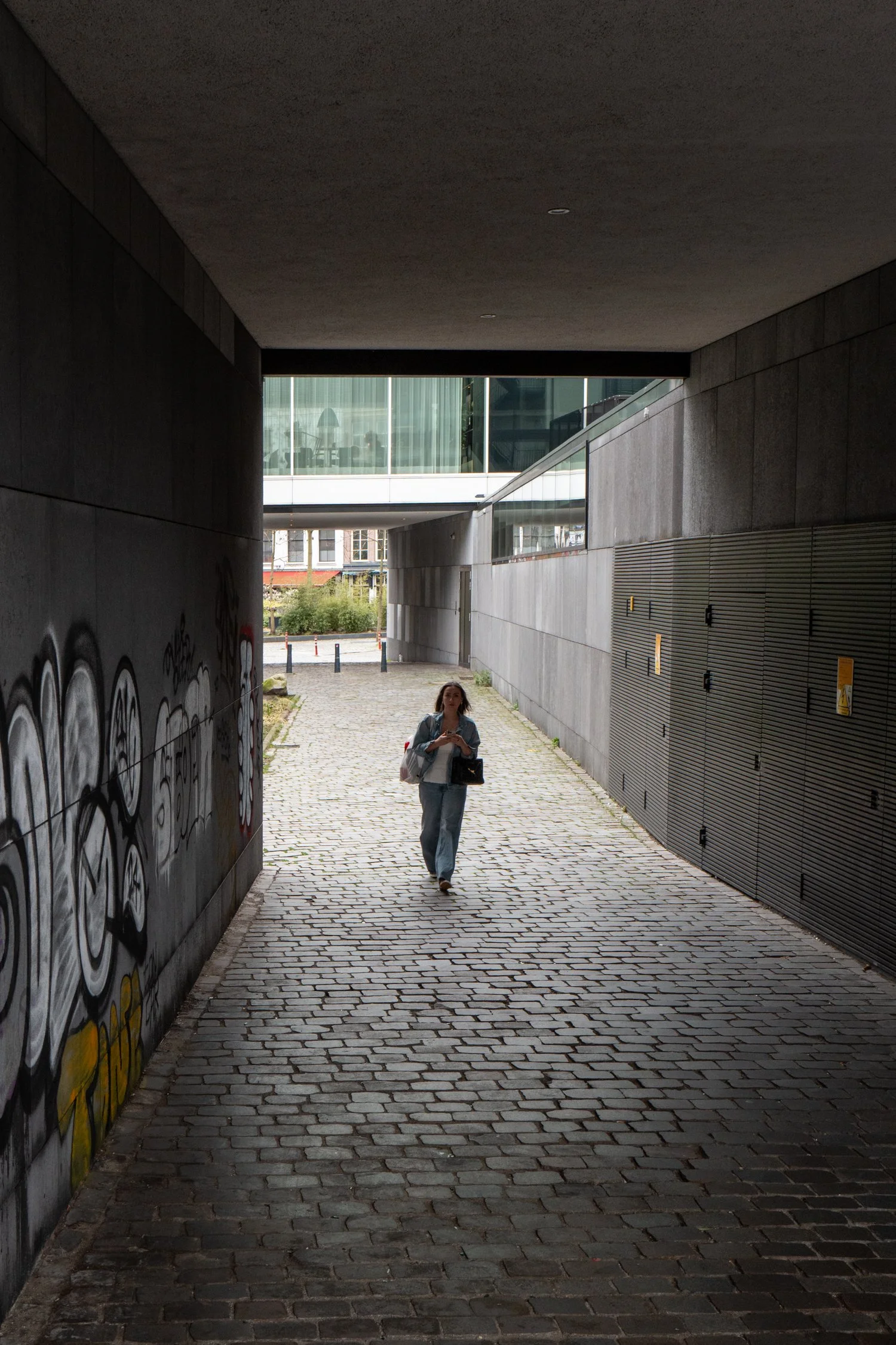 Une femme marche dans un passage souterrain pavé. À gauche, il y a un mur avec des graffitis. A droite, un mur en métal avec des boîtes électriques. À l'arrière, on voit l'extérieur avec des bâtiments et des plantes.