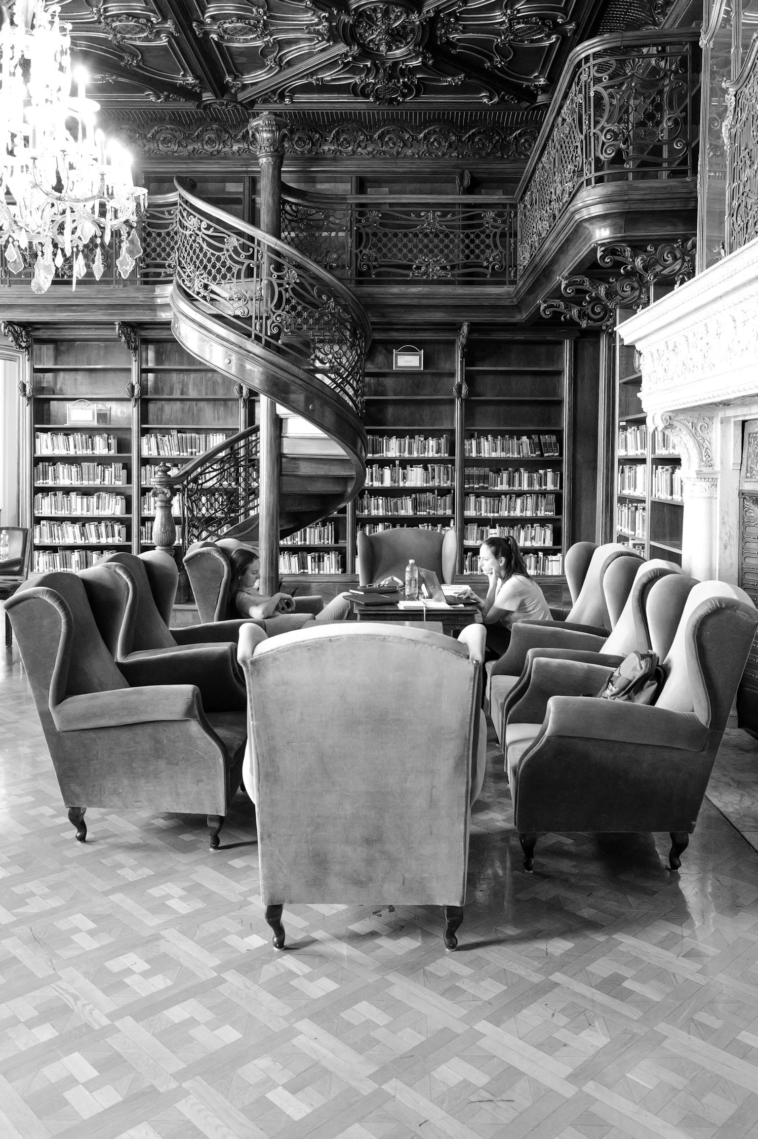 Une bibliothèque élégante avec des étagères remplies de livres en bois, un escalier en colimaçon en fer forgé, un chandelier en cristal suspendu au plafond orné de détails sculptés et deux femmes assises autour d'une table, travaillant sur un ordinat