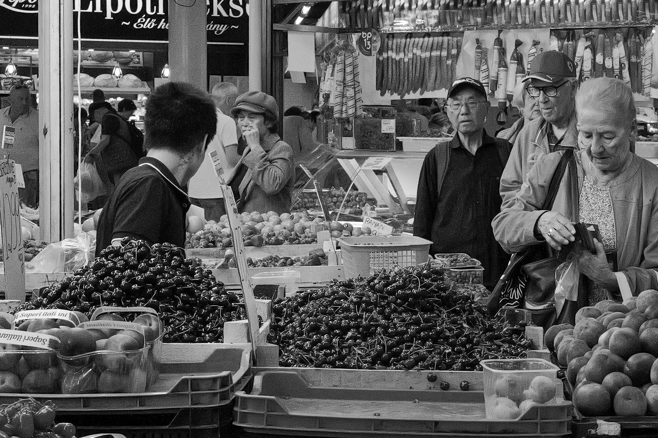 Marché avec plusieurs personnes regardant des fruits et légumes, étals remplis de cerises, pommes et autres fruits, décorations suspendues, enseigne en italien en arrière-plan.