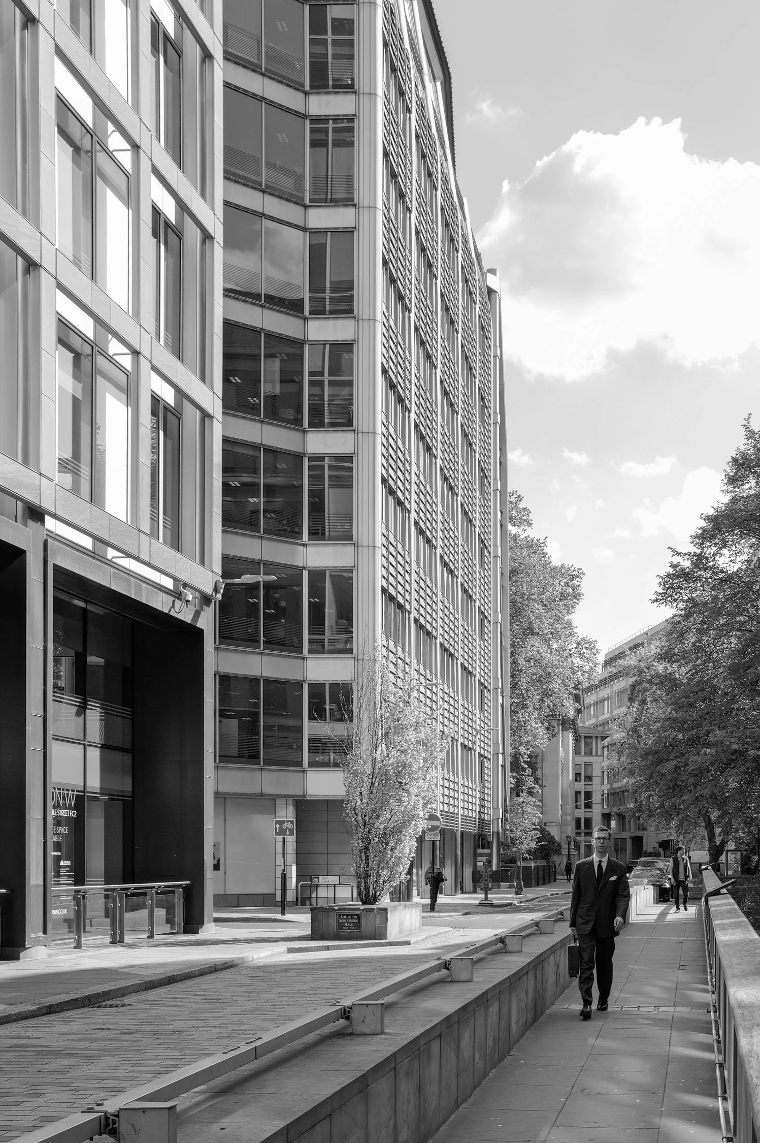 Une rue urbaine avec des immeubles modernes, des arbres et des piétons marchant dans la rue, en noir et blanc.