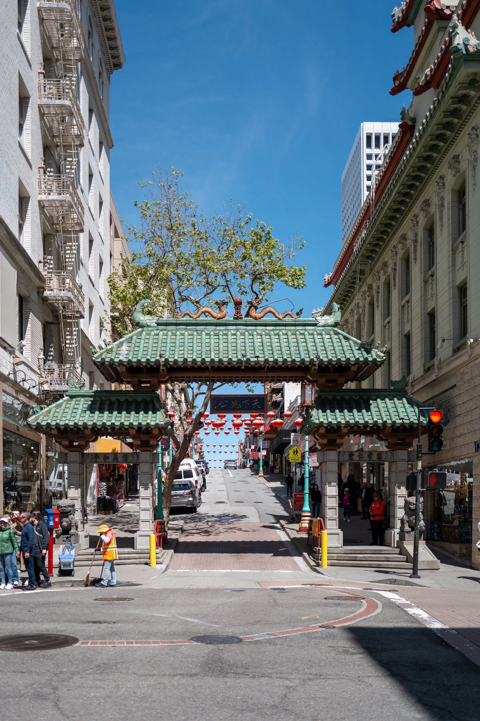 Une entrée d'inspiration chinoise avec un toit en tuiles vertes, des dragons décoratifs, des lanternes rouges suspendues, entourée d'immeubles modernes et de piétons dans une rue urbaine.