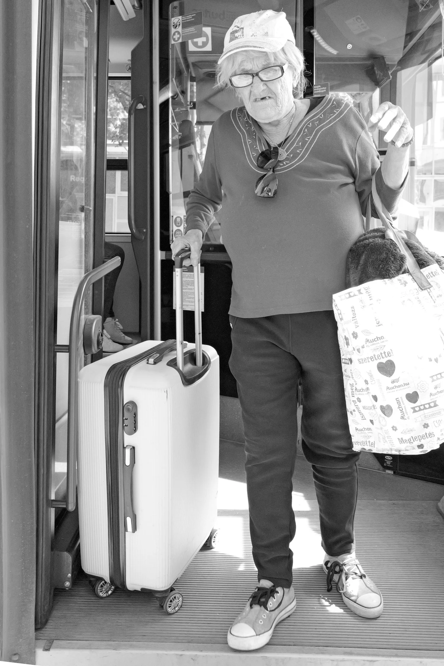 Une personne âgée portant une casquette, des lunettes, un sweat, des baskets, tenant une valise à roulettes et une grande sacoche, sortant d'un bus.